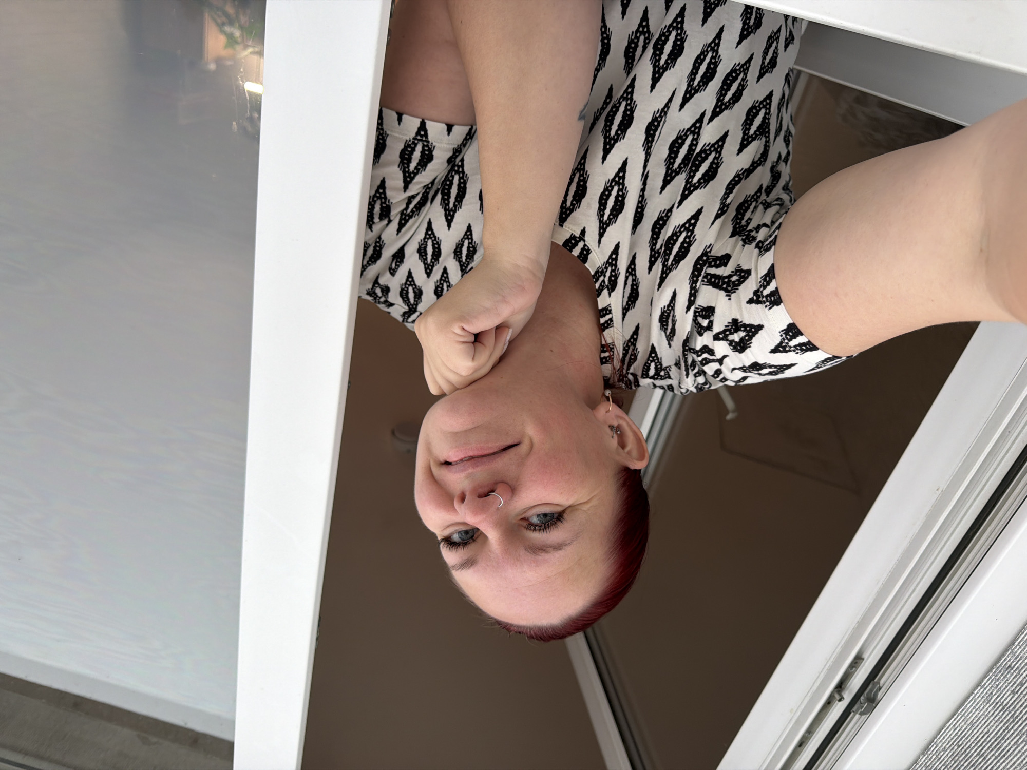 A woman with red hair and a nose ring is looking down at the camera. She is wearing a black and white patterned shirt. Her left arm is bent and her hand is near her chin. The setting appears to be inside near a white frame, possibly a window. The city of Chaikovsky, Russia, is not discernable within the image.