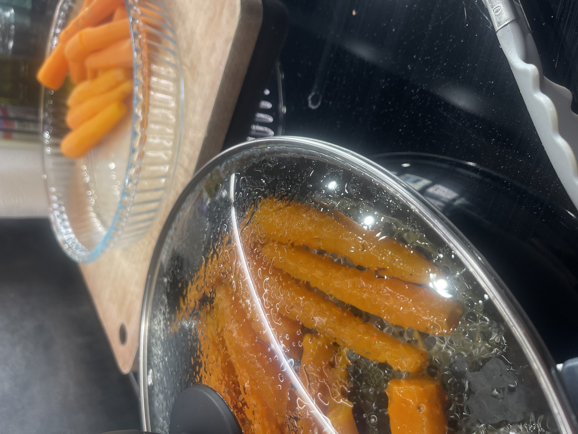 A close-up shot shows sliced carrots cooking in a pan covered by a glass lid. The carrots are bright orange and appear to be simmering in liquid, with small bubbles visible on the surface of the liquid and on the inside of the lid. To the right of the pan, a glass dish filled with more whole, raw carrots is visible, placed on a wooden cutting board. The scene is set on a black stovetop. There are no people visible in the image. The image does not provide any information about the location in the city of Karlovo, Bulgaria.