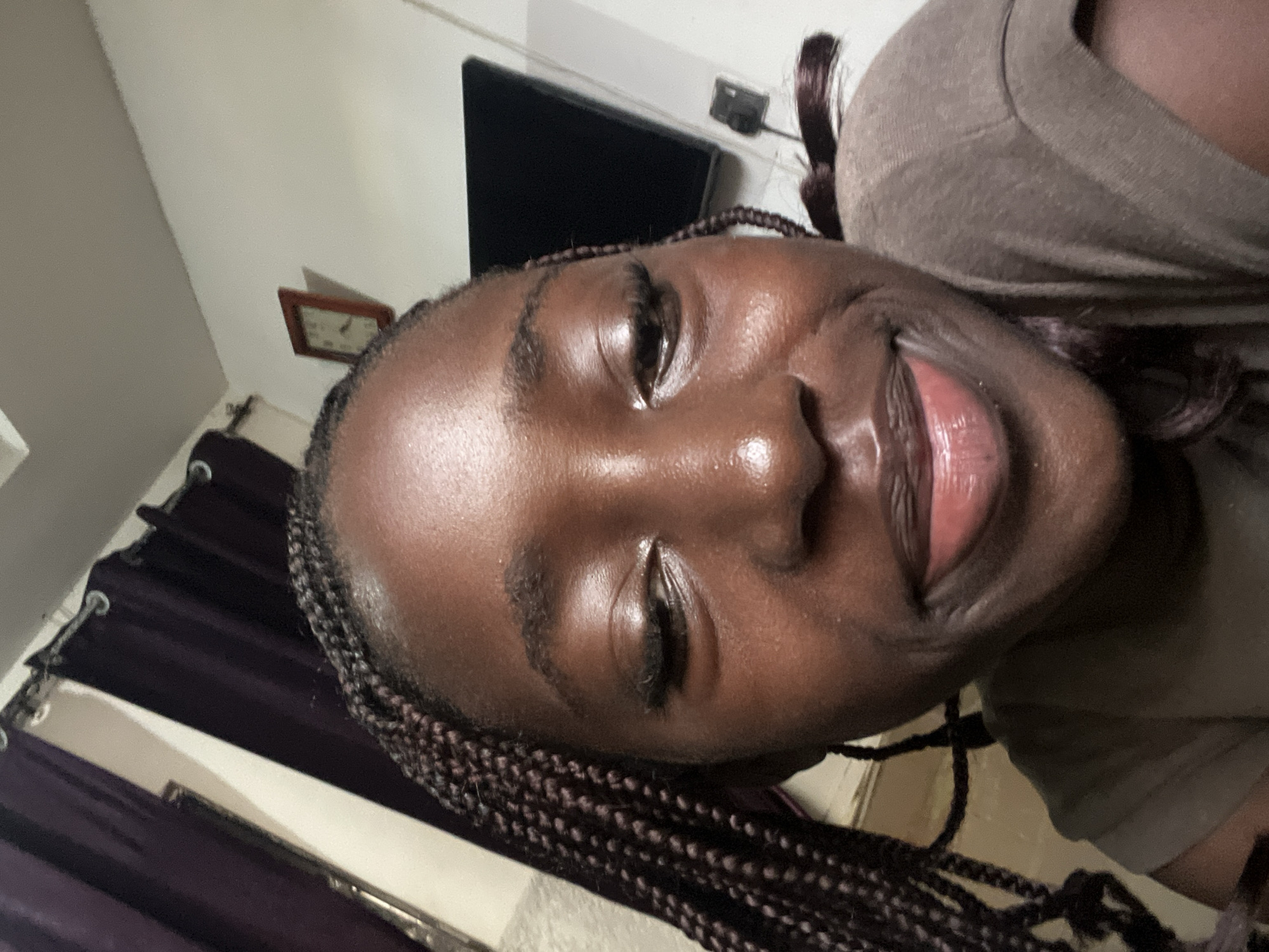 A close-up, slightly rotated portrait of a woman with dark skin. She has braided hair and is wearing a brown shirt. Her lips are slightly parted. In the background, there is a television screen and a clock on the wall. Purple curtains are visible to the left. The location is Abuja, Nigeria.