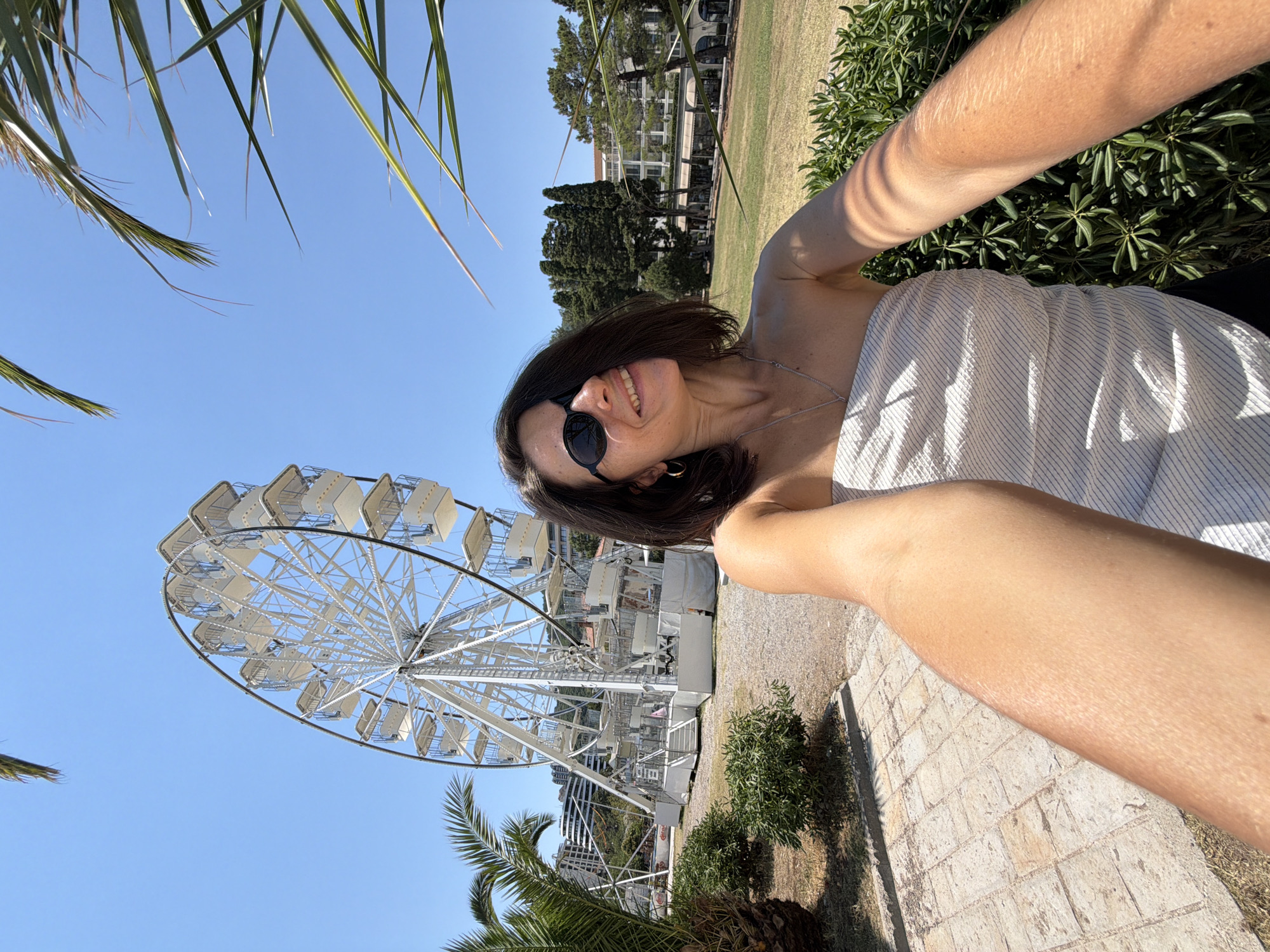 In this image, a woman with shoulder-length dark hair and sunglasses is captured in a selfie. She is wearing a strapless top and a necklace. She is standing outdoors, with a large white Ferris wheel visible in the background to her left. Palm trees and lush green bushes are present in the foreground and midground, along with a paved walkway. Behind the Ferris wheel, buildings and trees are visible, and the scene is set under a clear blue sky. The woman is smiling and looking towards the camera.