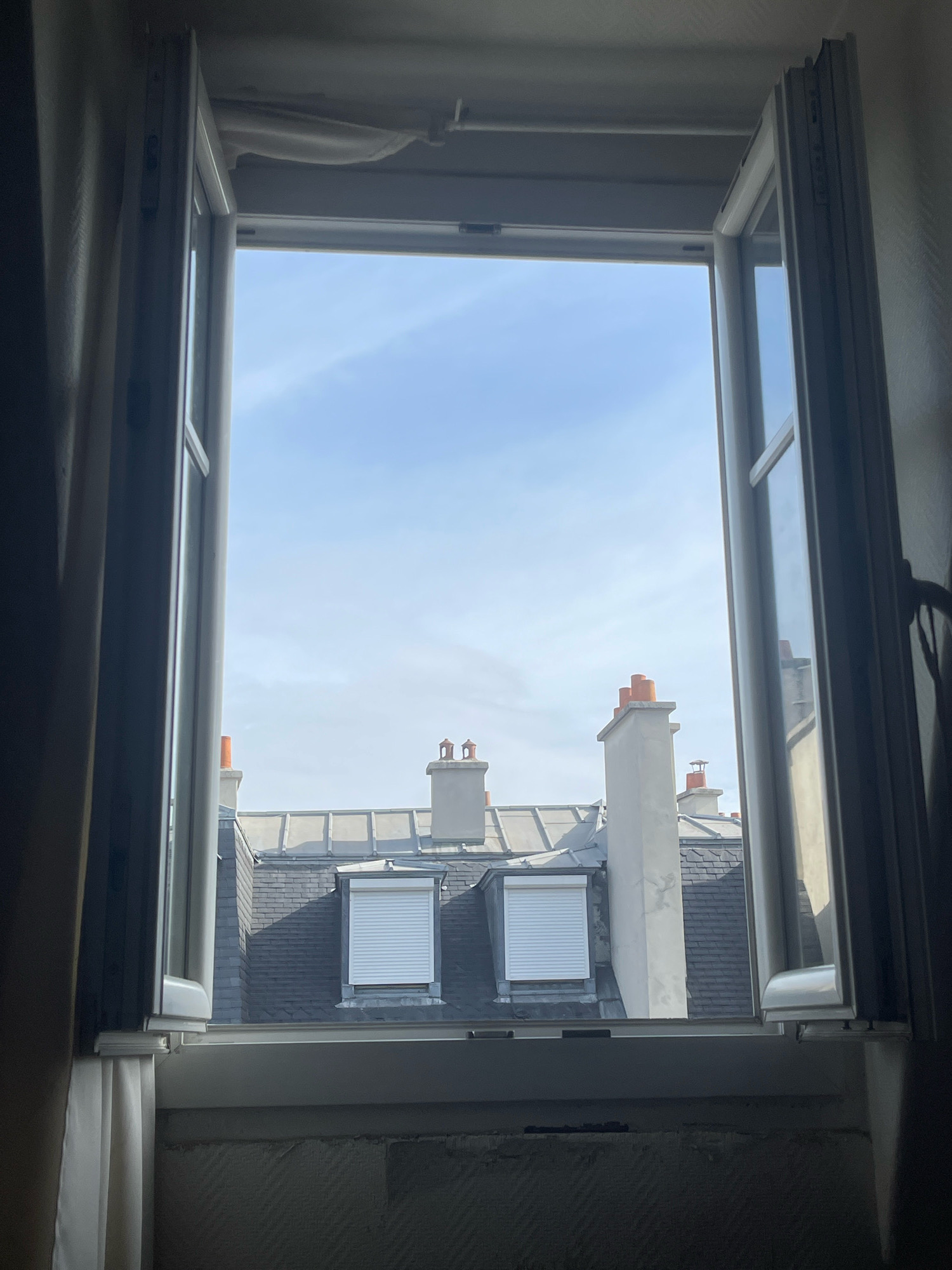 A wide-angle, high-angle, eye-level shot captures a view through an open window overlooking the rooftops of Paris, France. The window, with white frames, is open on both sides, revealing a clear blue sky with scattered clouds.
Dominating the middle ground are several slate-tiled rooftops, punctuated by traditional Parisian chimneys with orange caps. Some rooftops feature dormer windows with white roll-up shutters. The architecture suggests a classic Haussmannian style common in Paris. The light is soft and diffused, indicating it is likely daytime, possibly morning or late afternoon.
In the foreground, the interior of the room is visible. A beige curtain hangs on the left side of the window. The windowsill is textured, and below it, a roughly textured wall is present. The overall impression is one of a serene, urban landscape viewed from a comfortable vantage point within a Parisian apartment.