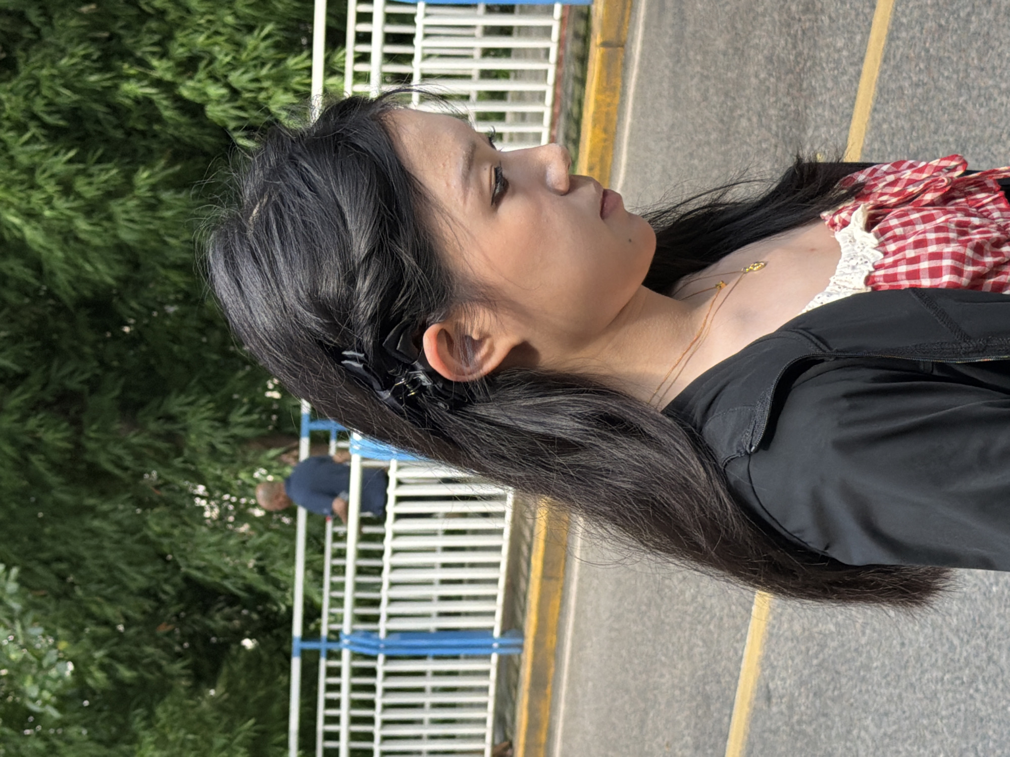 A medium shot shows a young Asian woman in profile, facing right and looking down. She has long, dark hair secured with a black clip, and she is wearing a black jacket over a red and white gingham top with white lace trim. A thin gold chain with two small pendants rests on her collarbone. She has light skin, dark eyes, and her expression is neutral. The background is blurred, but a white railing with blue vertical supports is visible behind her, suggesting she is on a bridge or elevated walkway. Beyond the railing, lush green trees form a dense backdrop, and two figures, one in a dark blue shirt and another in a lighter shirt, are faintly visible in the distance. The ground beneath her appears to be asphalt, with a yellow line marking the edge of a roadway. The scene is outdoors, under bright sunlight. The location is identified as Yuzhong District, China.