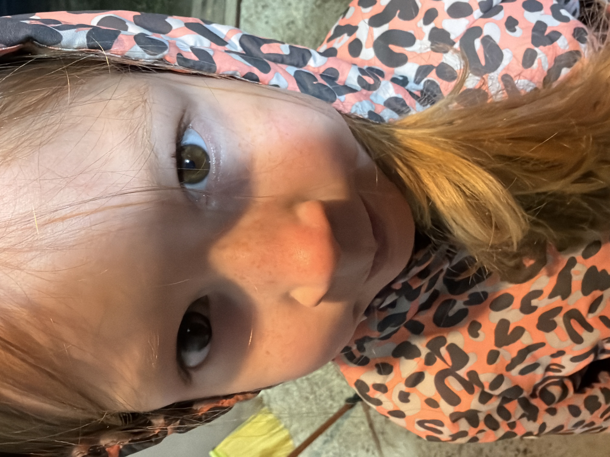 This close-up portrait, taken in Markovo, Russia, features a young child looking directly at the viewer. The child has fair skin, visible freckles on the nose and cheeks, and expressive brown eyes. Light brown or blonde hair frames the child's face, with some strands falling across the forehead. The child is wearing a hooded garment, likely a jacket, featuring a distinctive leopard print pattern in shades of coral/peach, grey, and black. The lighting is soft and even, suggesting an indoor setting or a shaded outdoor area. The background is blurred and indistinct, showing hints of a yellow object and a textured surface towards the bottom. The child's expression appears neutral or slightly curious. There is no visible text in the image.