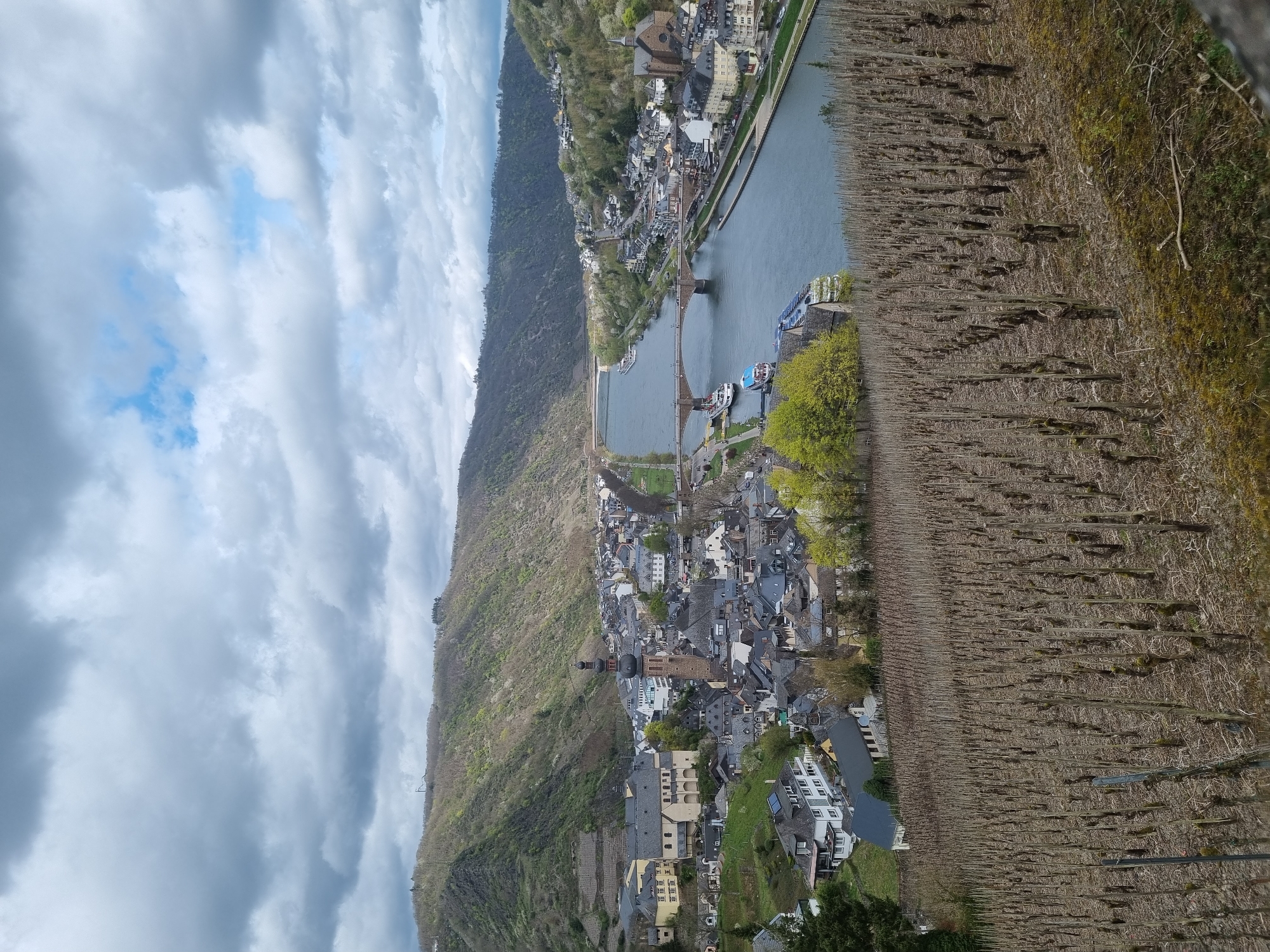 The image shows an elevated, slightly oblique view of Cochem, Germany, situated along the Moselle River. The sky is partially cloudy with patches of blue visible. On the left side of the frame, a wooded hillside rises steeply. The Moselle River snakes through the scene, and a bridge connects the two sides of the town. Buildings with dark roofs, typical of the region, cluster along the riverbank and extend up the slope. A prominent church steeple is visible among the buildings. On the right, vineyard terraces are visible. The vines are bare, indicating a dormant or early stage of growth. The overall impression is of a picturesque German town nestled in a valley landscape.