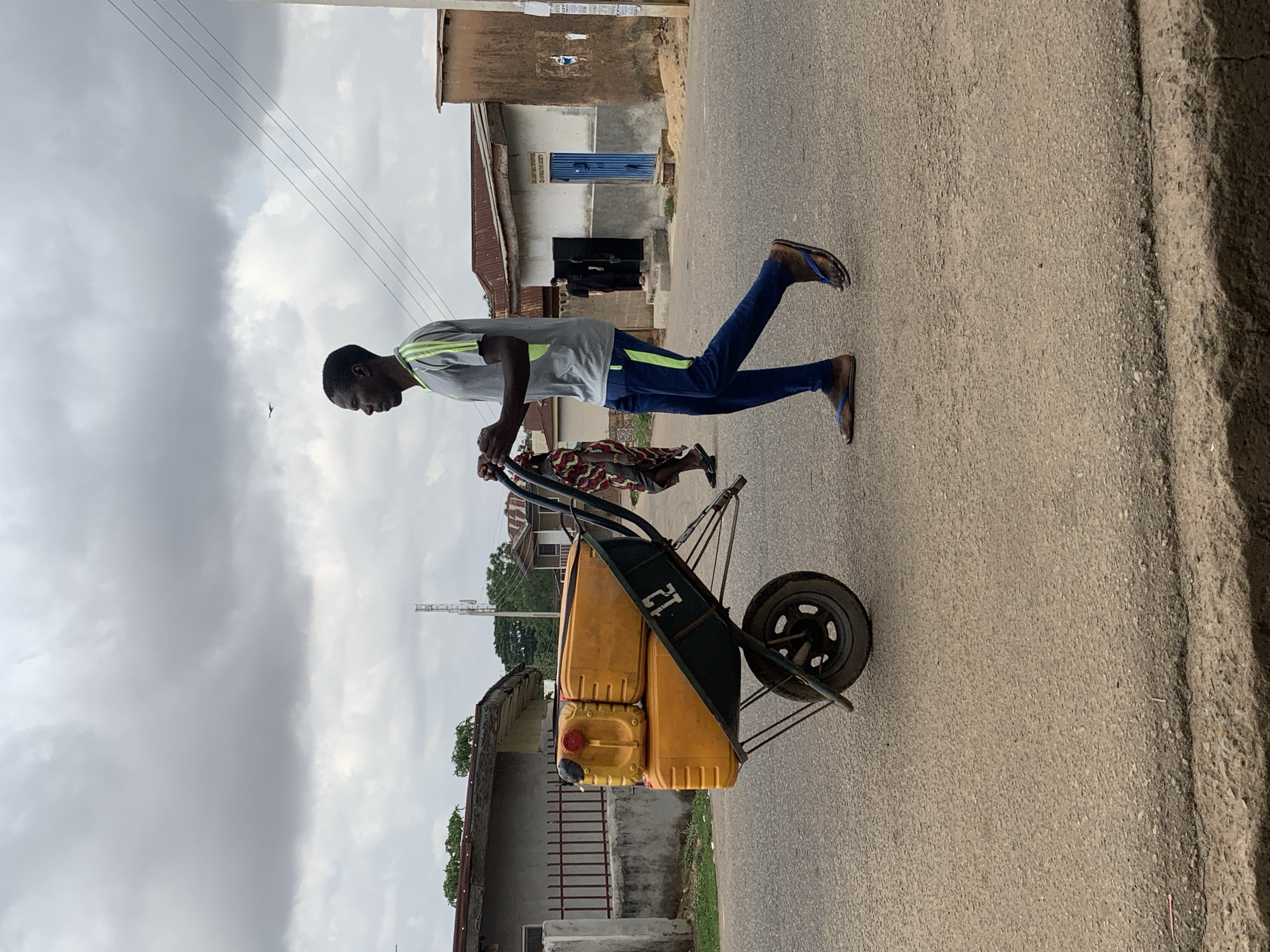 A man is walking down a paved road in Gura Top, Nigeria, pushing a wheelbarrow. The wheelbarrow contains three yellow plastic jerrycans. The man is wearing a gray t-shirt with green stripes on the shoulders, blue sweatpants, and flip-flops. There are buildings on both sides of the road, and a cloudy sky is overhead. A person walks on the right behind the wheelbarrow. A cell tower can be seen in the distance.
