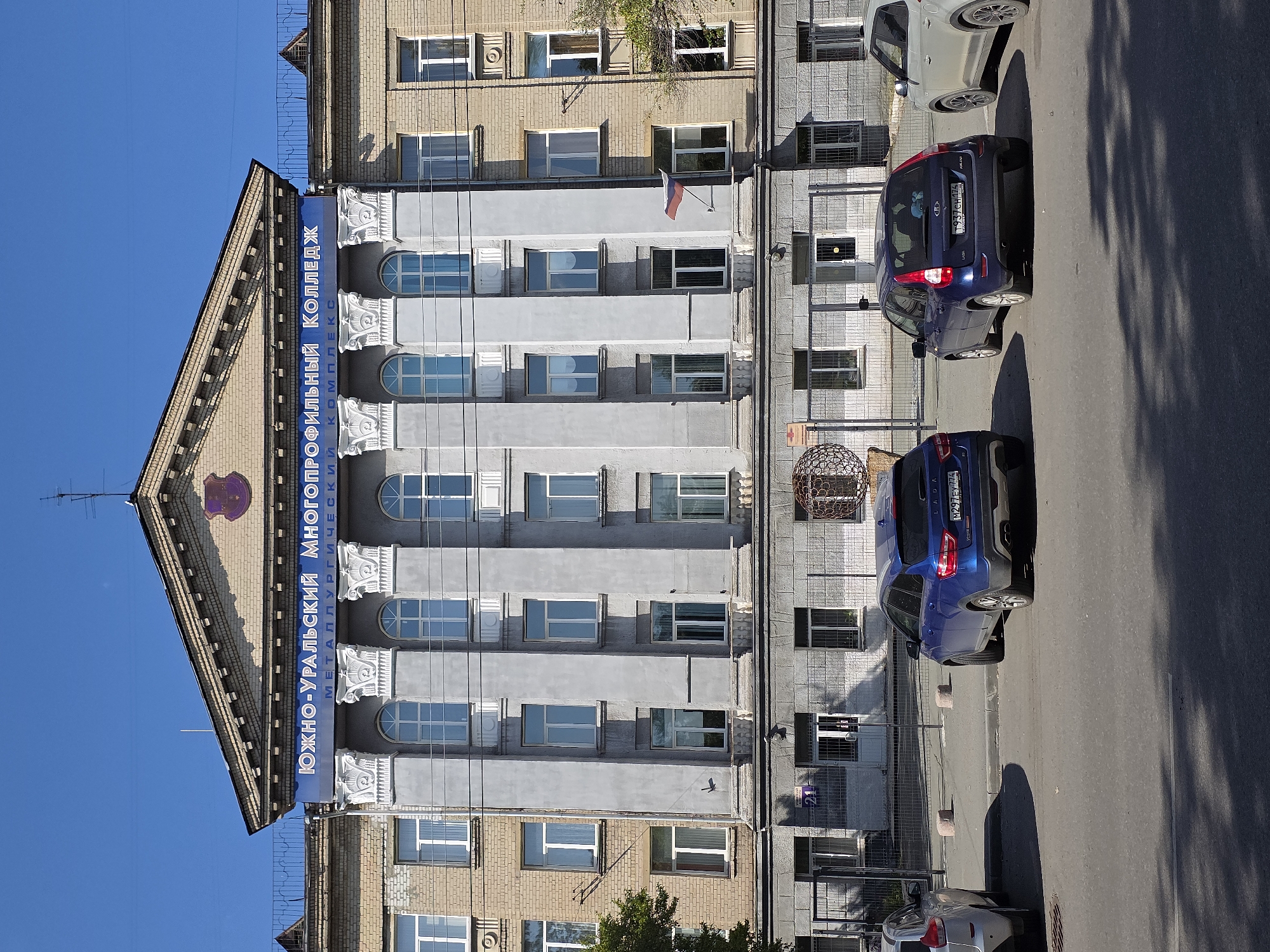 The image shows the exterior of the Yuzhno-Uralsky Multidisciplinary College in Chelyabinsk, Russia. The building is a multi-story structure with a classical facade, featuring large columns and ornate detailing. The college's name is displayed prominently in large blue lettering across the top of the building. The lower portion of the building has a gray stone facade with multiple windows and doors. In front of the building, there is a parking lot with several cars. Two blue cars are parked in the foreground, and a silver car is parked to the right. The scene is under a clear blue sky on a sunny day. The overall impression is one of a well-maintained educational institution.