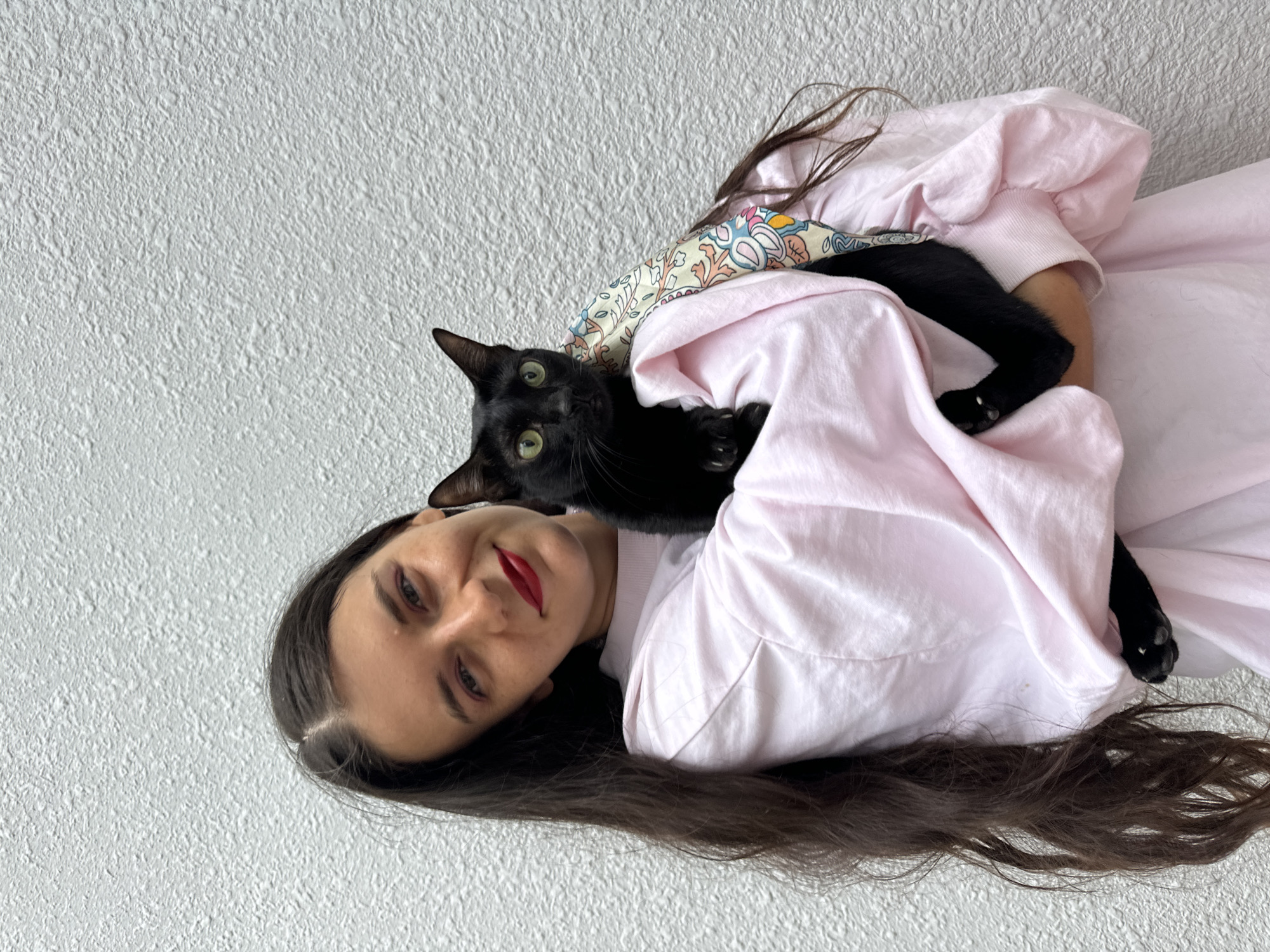A young woman with long dark hair and red lipstick is holding a black cat. She is wearing a light pink sweatshirt. The cat has bright green eyes and is looking towards the camera. A patterned fabric is visible around the cat. They are standing in front of a textured white wall. The setting appears to be outdoors, possibly in the city of Mũi Né, Vietnam, given the light and the whitewashed wall commonly found in coastal areas. The woman is smiling, and the cat seems relaxed in her arms. The overall scene is casual and affectionate.