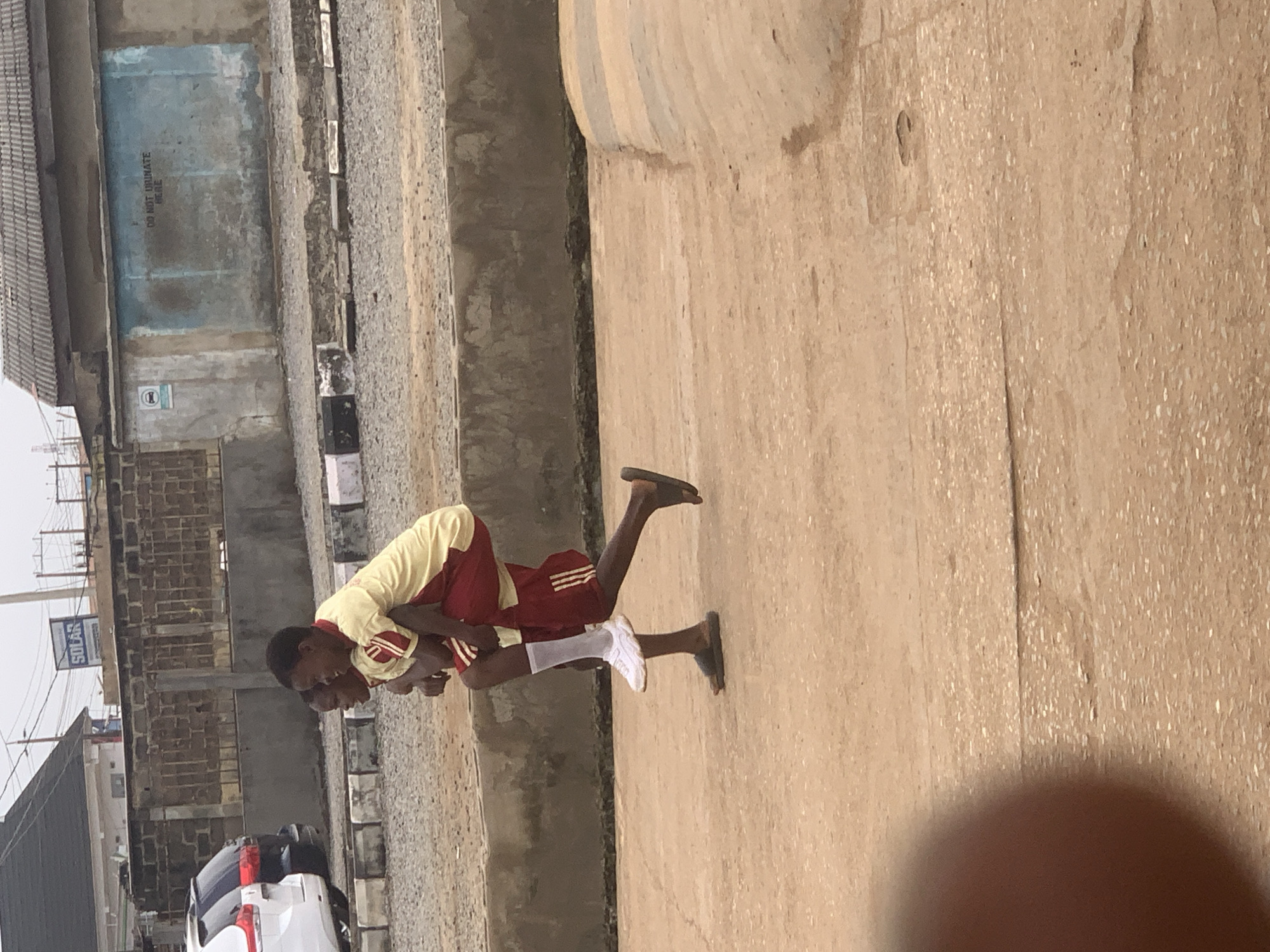 In the city of Shagamu, Nigeria, two young boys are captured in a side-view, full-body shot as one carries the other on his back, walking on a paved street. The boy being carried, wearing a yellow and red short-sleeved shirt with red shorts, has his arms around the carrier's neck. The carrier, dressed in a similar yellow and red outfit, sports red shorts and white socks with white sneakers, while his bare feet are shod in dark flip-flops. They are moving from left to right across the frame. The background features a grey stone wall topped with a dark fence, behind which are buildings, including one with a "SOLAR" sign. Further in the background, a light blue wall displays the words "DO NOT URINATE HERE" in black lettering, accompanied by a circular sign with a white figure inside. A section of a white car and a black vehicle are visible on the left side of the image, parked behind a low barrier. The ground is predominantly a reddish-brown concrete surface, with a section of darker, rougher terrain in the mid-ground. The lighting suggests an overcast day.