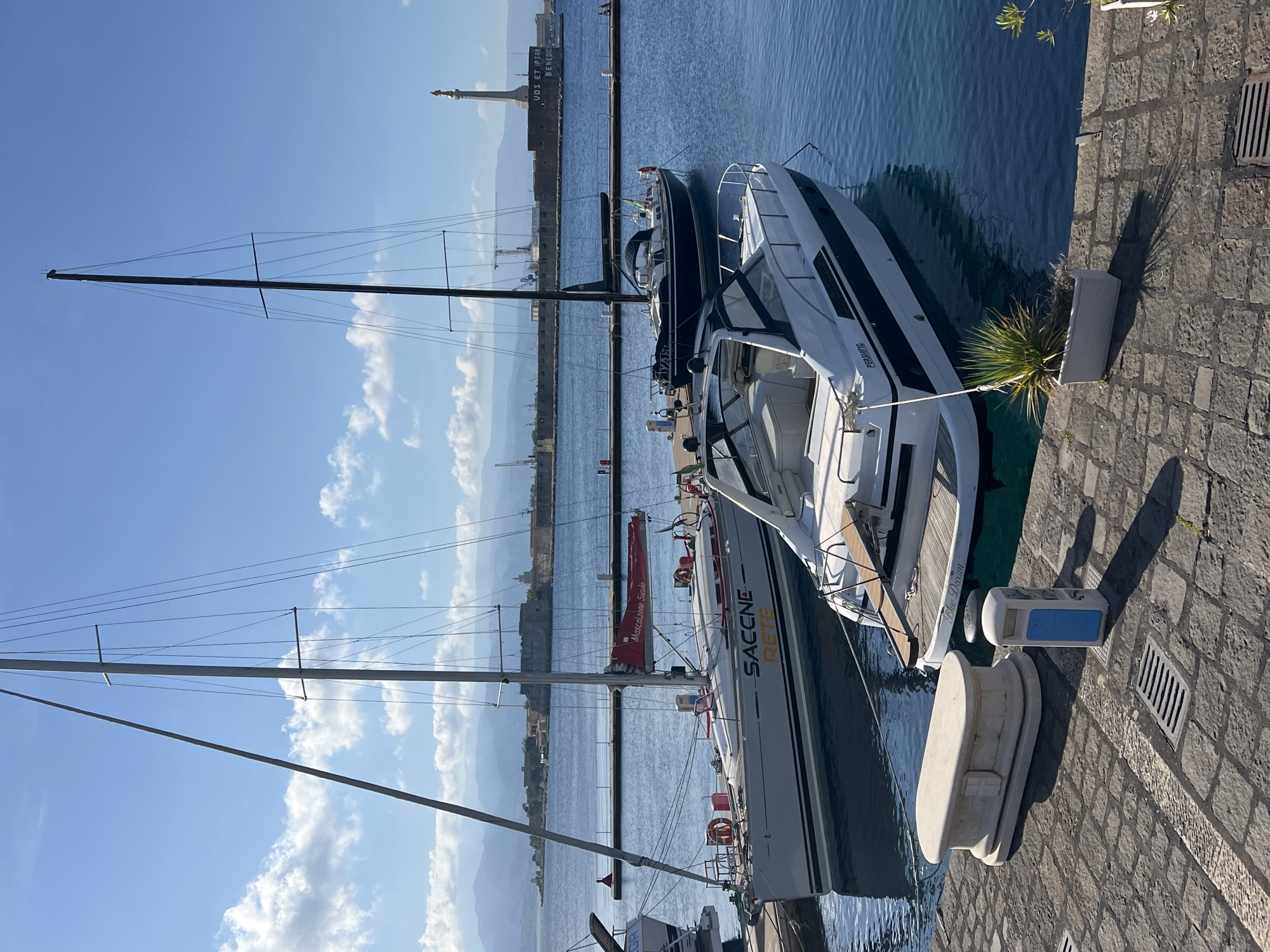 The image presents a daytime view of a marina in Messina, Italy, under a bright blue sky with scattered white cumulus clouds.
In the foreground, a cobblestone quay features a large, light-colored stone bollard with an embossed cross-like design, positioned left of center. Adjacent to it on the right is a shorter, grey utility post with a blue top. A rectangular metal drain grating is set into the cobblestones in the lower-left quadrant. At the quay's edge on the lower right, a white planter holds green, spiky foliage.
Three boats are moored in the calm, blue water. The closest boat, positioned in the midground on the right, is a white and black motor yacht with "PERSHING" visible on its hull and "Blue Dream" on its stern. It has a light-colored swim platform at its stern. To the left of the motor yacht, a grey monohull sailboat with black hull stripes is moored to a floating brown dock. This sailboat displays "SACCNE" and "RETE" in orange lettering on its hull, and "Mascalzone Siculo" on its red mainsail cover. Behind the motor yacht, a dark blue sailboat with a black mast and dark blue sail cover is also moored to a similar floating dock.
In the background, across the blue water, a long grey breakwater is prominent. On its far-right end, a tall column supports the "Madonnina del Porto" (Madonna della Lettera) statue, with the Latin inscription "VOS ET IPSAM BENEDICIMUS" clearly visible on its base. To the left of this landmark, a large, grey, historic fortress (Forte San Salvatore) stands at the harbor entrance. Further along the distant shoreline, industrial cranes and port structures are discernible. A range of hazy blue mountains forms the horizon. Strong shadows cast by objects in the foreground indicate bright sunlight from the left. No individuals are clearly visible in the foreground or midground.