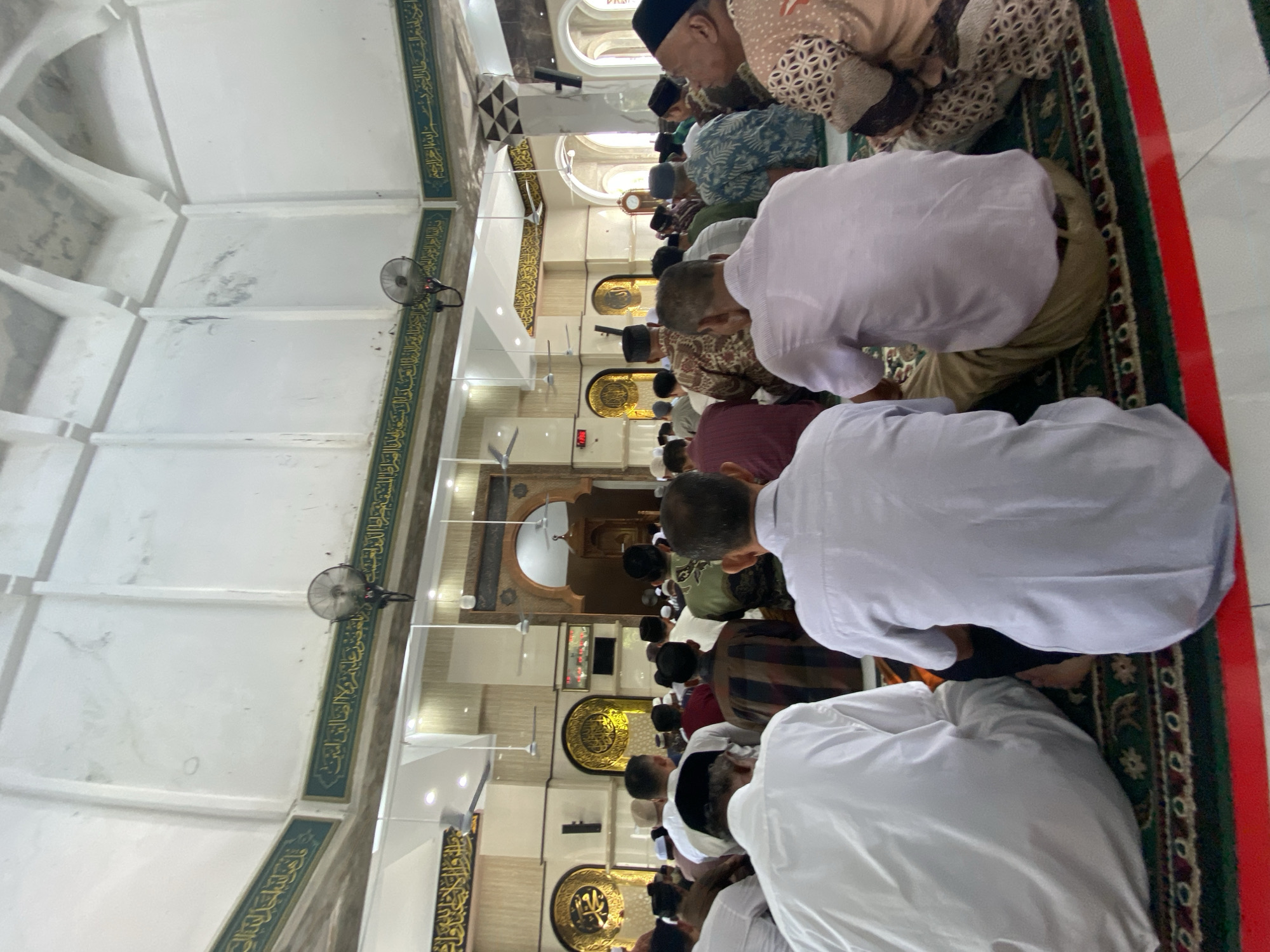 This image depicts a group of Muslim men seated on prayer rugs inside a mosque, facing towards an altar area. The men are seen from behind, dressed in traditional Islamic attire, including various patterns and solid colors. The mosque's interior features high ceilings with arched structures and decorative Islamic calligraphy along the walls and ceiling. Ceiling fans and lighting fixtures are visible. The floor is covered with a patterned prayer rug. The altar area appears to have a wooden structure and ornate gold decorations. The overall scene suggests a religious gathering or prayer service. This image is of the Baiturrahman Grand Mosque in Lhokseumawe, Indonesia.