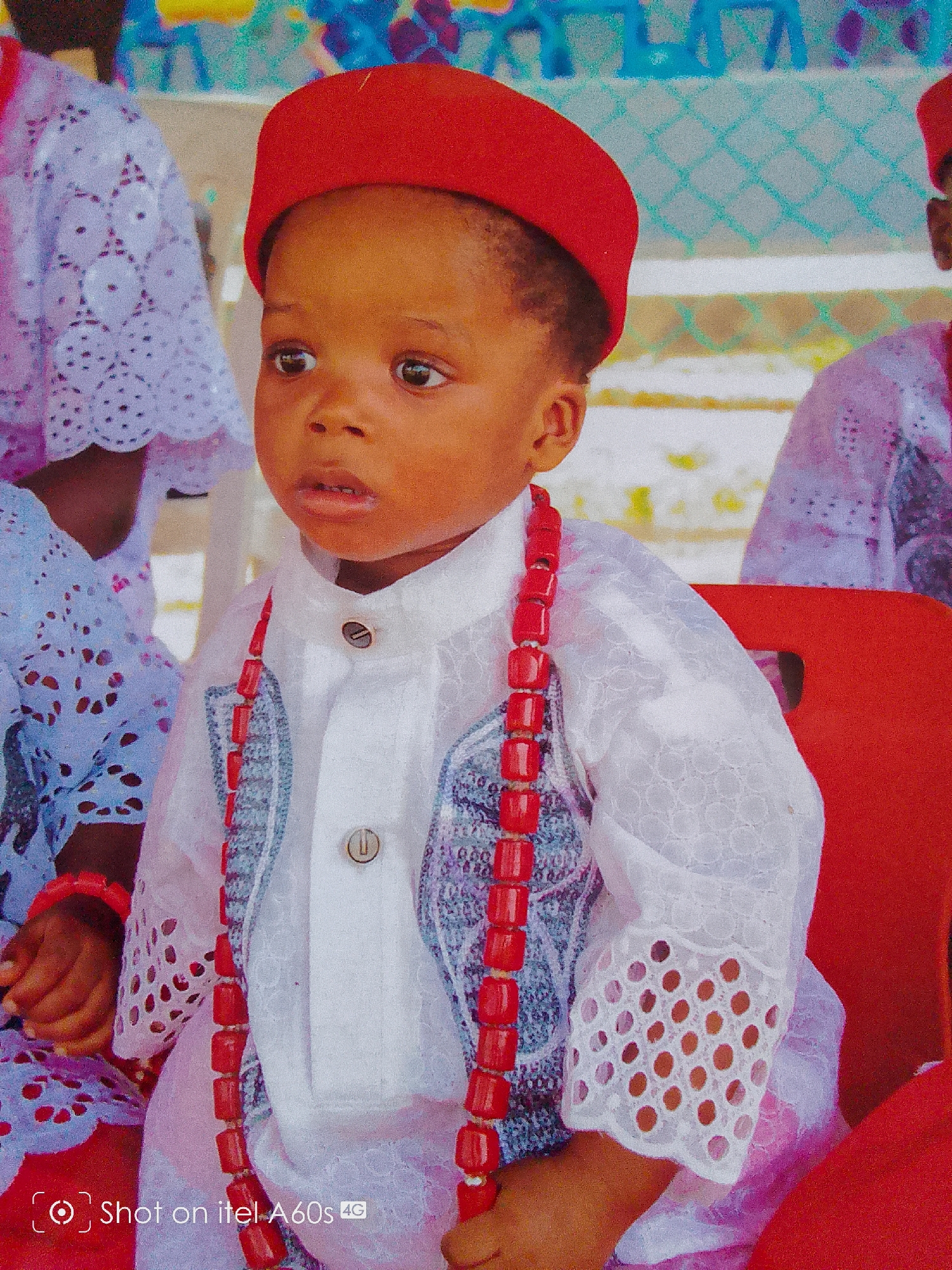 A young child sits, adorned in a red cap and necklace, wearing a white and patterned traditional garment. The child is the primary subject of the image, appearing to be at an event or gathering. Other individuals are partially visible in the background, also dressed in patterned attire. The background shows hints of a fence or railing, and chairs. The image was taken with an itel A60s device. There is no specific detail to identify the exact location in Idimu, Nigeria.