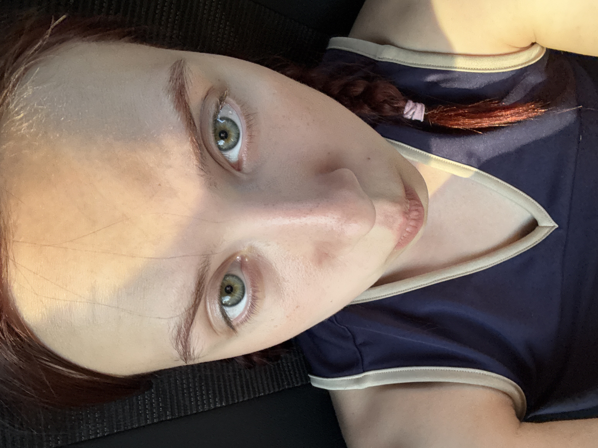 A close-up, rotated portrait of a fair-skinned person with reddish-brown hair and green eyes, likely female, wearing a dark blue garment with light trim. The person is looking upwards towards the camera with a slight sideways tilt. A small braid is visible near the neckline of the garment. The background appears dark and undefined. There is no indication of the location being Bol'shoe Frolovo, Russia in the image.