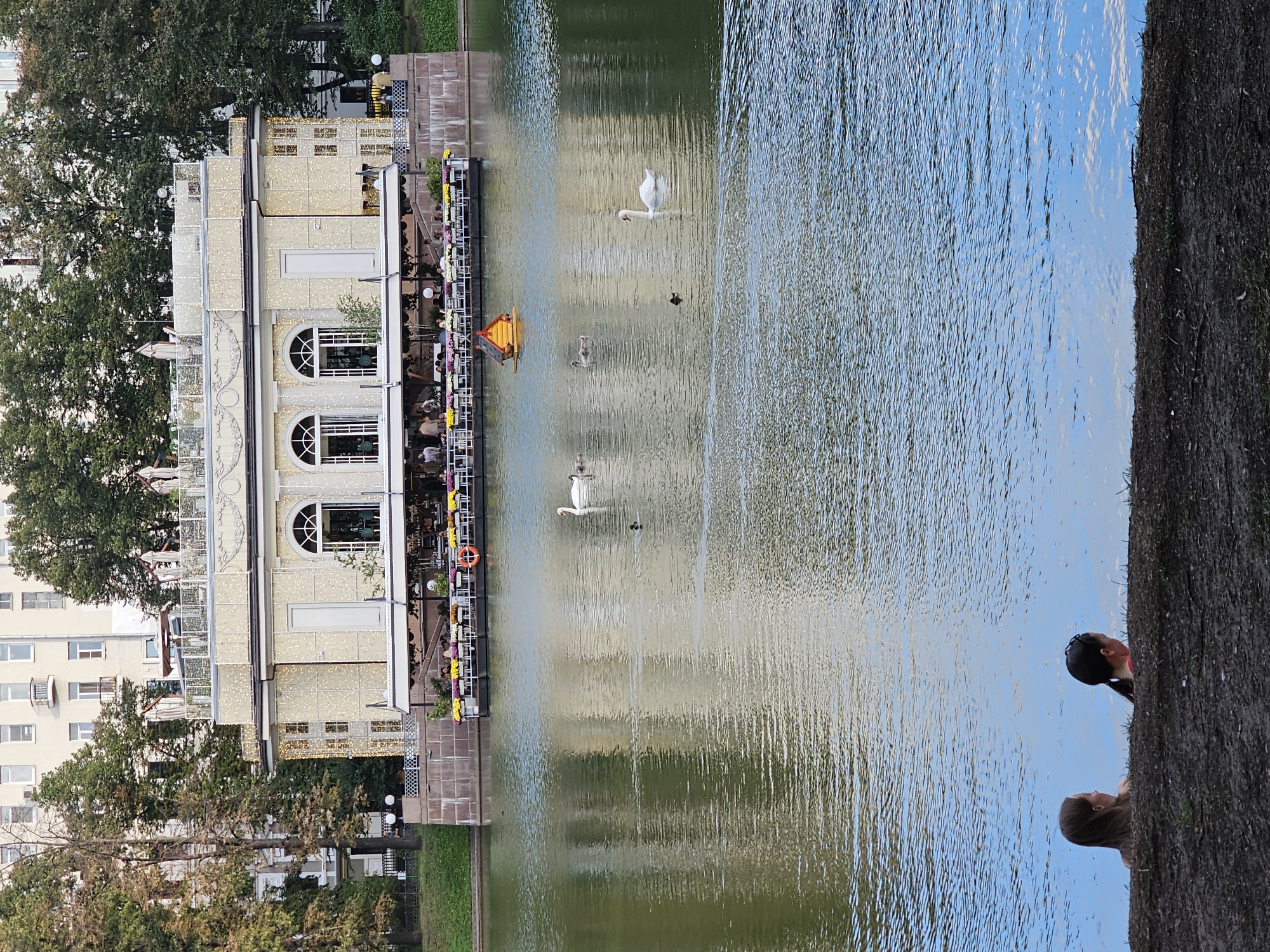 This image depicts a scenic view of a pond in Moscow, Russia. In the foreground, two young women are partially submerged in the water, with their backs to the camera, looking out at the pond. The pond is calm, with ripples on the surface reflecting the surrounding environment. Several swans and ducks are visible on the water. In the background, a large, ornate building stands beside the pond. The building appears to be a restaurant or cafe, with outdoor seating and a decorative facade illuminated by numerous small lights. Beyond the building, trees and other structures are visible, suggesting an urban park setting. The overall atmosphere is serene and picturesque, capturing a moment of leisure in a beautiful natural environment within the city.