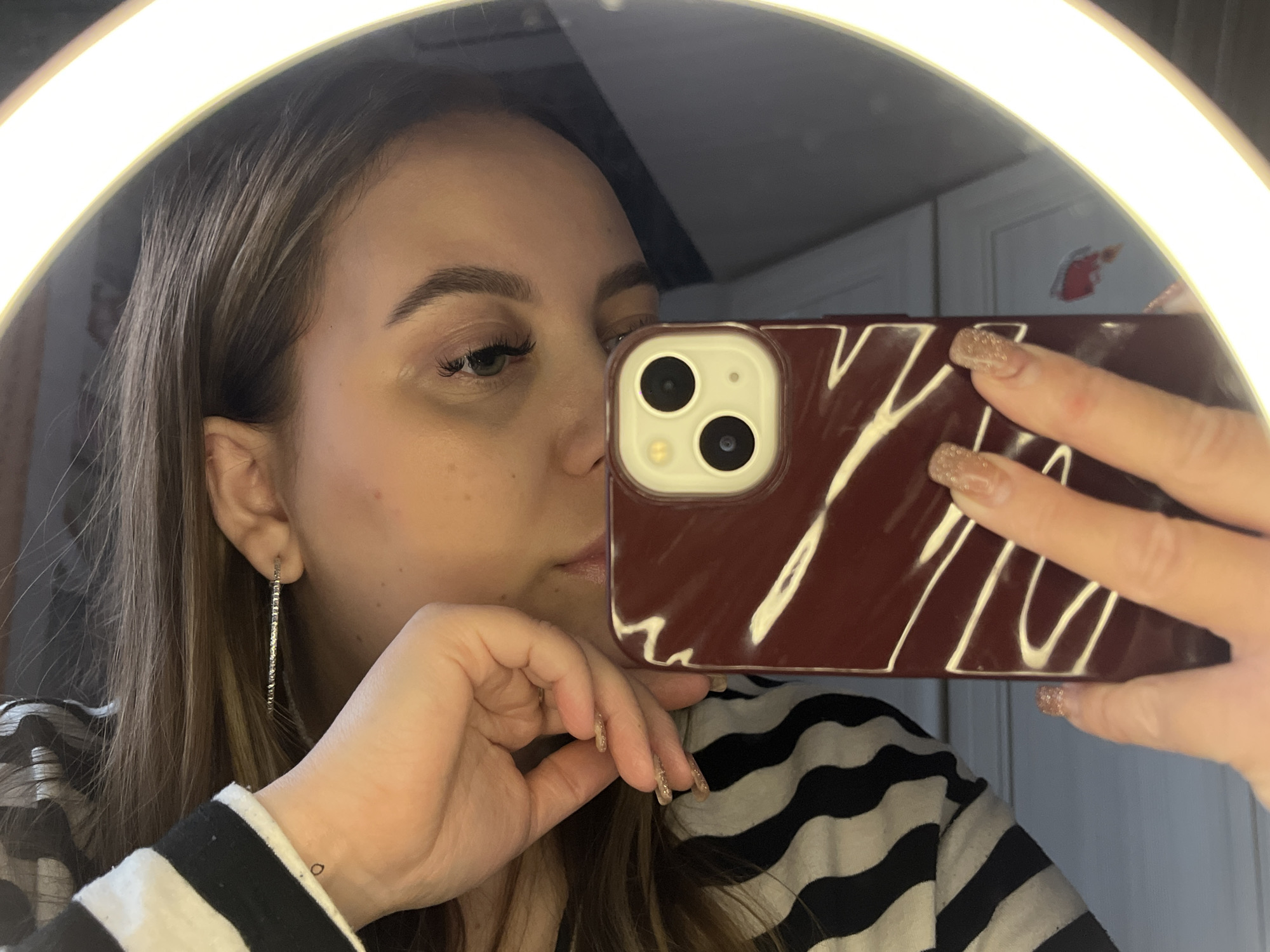 A woman is taking a selfie in a mirror. The mirror is illuminated by a ring light. She is holding a phone with a maroon case that has white stripes or patterns on it. The woman has light skin, brown hair, and is wearing long, thin, silver earrings. She has short, glittery nail polish. The woman is wearing a black and white striped shirt. The background includes a wall with a sticker or picture of a red character. It is not possible to determine if the location is Novopokrovskaya, Russia, based solely on the visual information provided in the image.