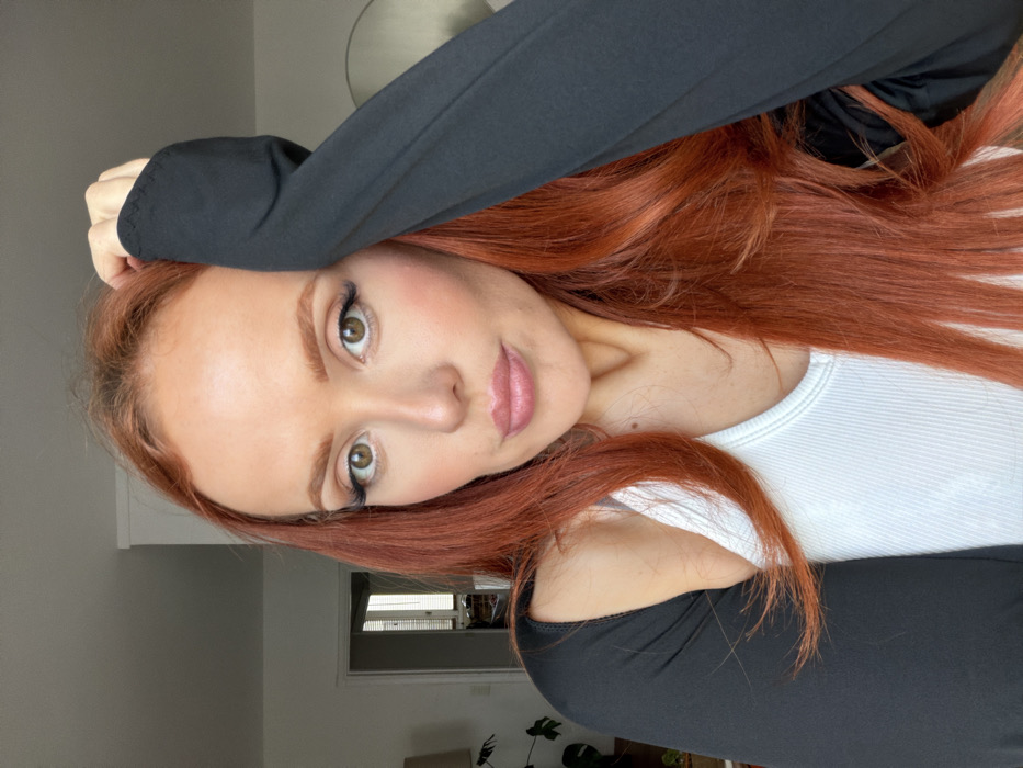 The image depicts an adult female in a close-up, indoor setting. She possesses long, wavy auburn hair, fair skin, and green eyes accentuated with dark eyeliner and eyelashes. Her lips are lightly colored with pink lipstick. She wears a white ribbed tank top partially obscured by a black garment that covers her arms and shoulders. Her right hand rests on her head, with fingers intermingled with her hair. Her gaze is directed towards the camera. The background consists of light-colored walls in what appears to be a residential interior. A doorway on the left reveals a secondary room with a visible lamp, a potted plant, and a window fitted with vertical blinds. A curvilinear object, possibly a mirror or wall hanging, is partially visible on the right wall. There are no discernible architectural or environmental elements within the frame that identify the specific location as Paris, France.