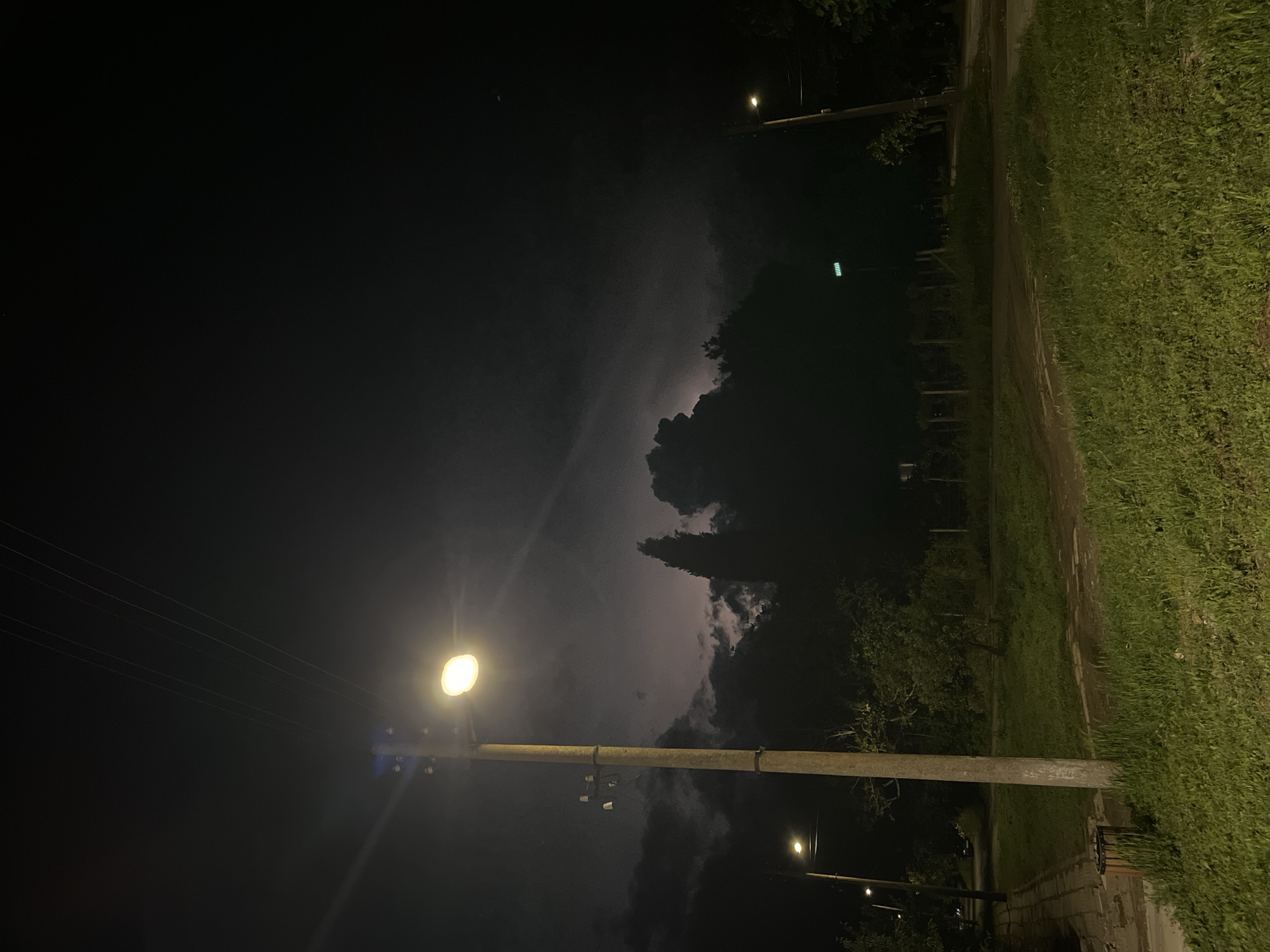 The image shows a night scene in Lozarevo, Bulgaria. A street is visible, with grass on the right side. Two utility poles with streetlights illuminate the area. In the background, dark clouds are present, with a possible lightning strike visible. The left side of the image is dark, but utility lines can be seen connecting to the lower utility pole.