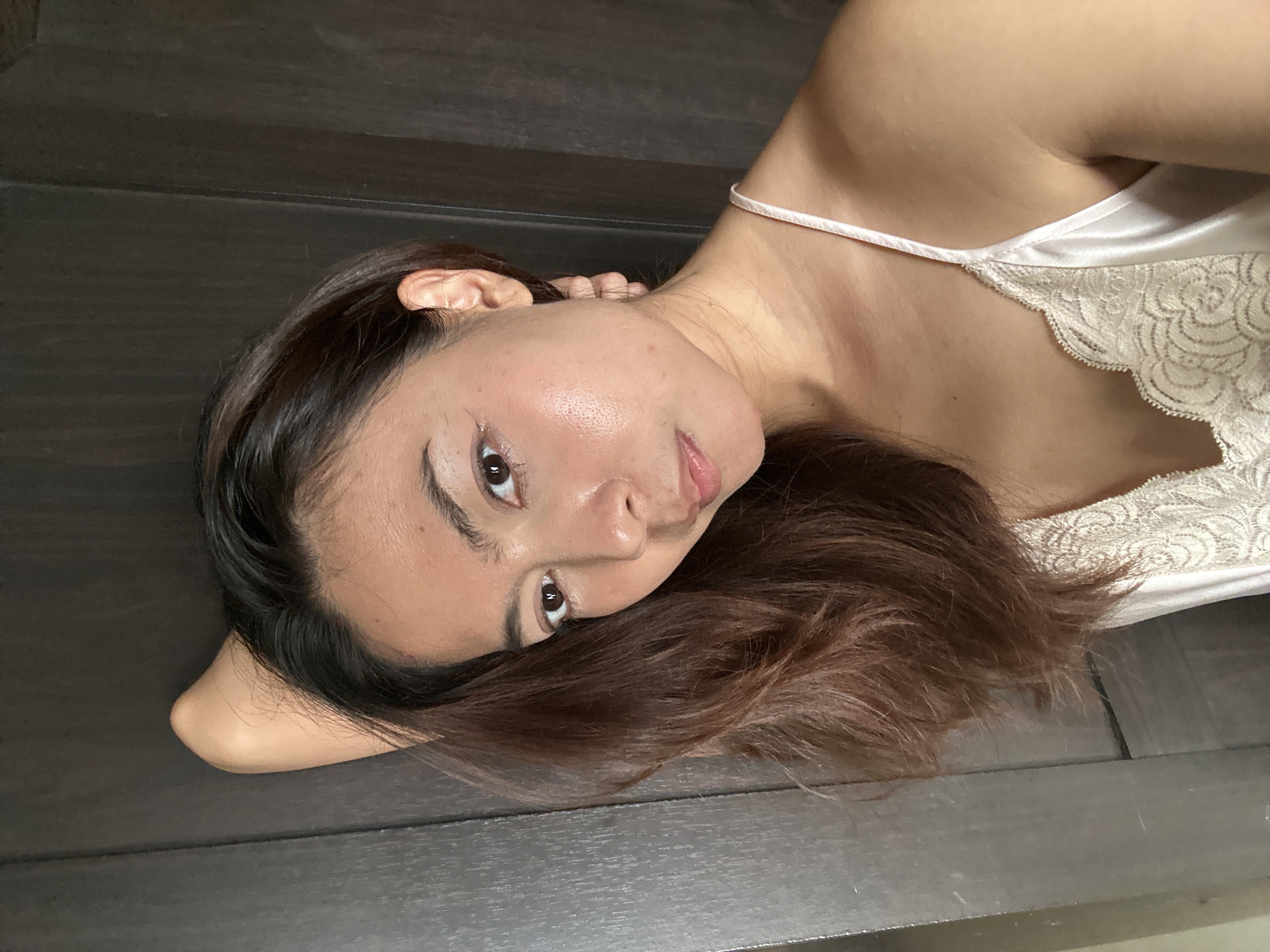 A medium shot captures a woman from the chest up, positioned to the left of the frame. She has long, dark brown hair that cascades over her left shoulder and is partially obscuring her face. Her right arm is bent, with her hand resting behind her head, accentuating the curve of her upper arm and shoulder. She is wearing a light-colored, silky camisole with delicate lace trim along the neckline. The background appears to be a dark, textured wall, possibly wood paneling, which provides a stark contrast to her skin tone and attire. The lighting is soft and diffused, highlighting the contours of her face and creating a subtle sheen on her skin. There is no clear indication of her location in Khlong Luang Town Municipality, Thailand, as the background is generic and lacks any identifying landmarks or architectural features specific to the region.