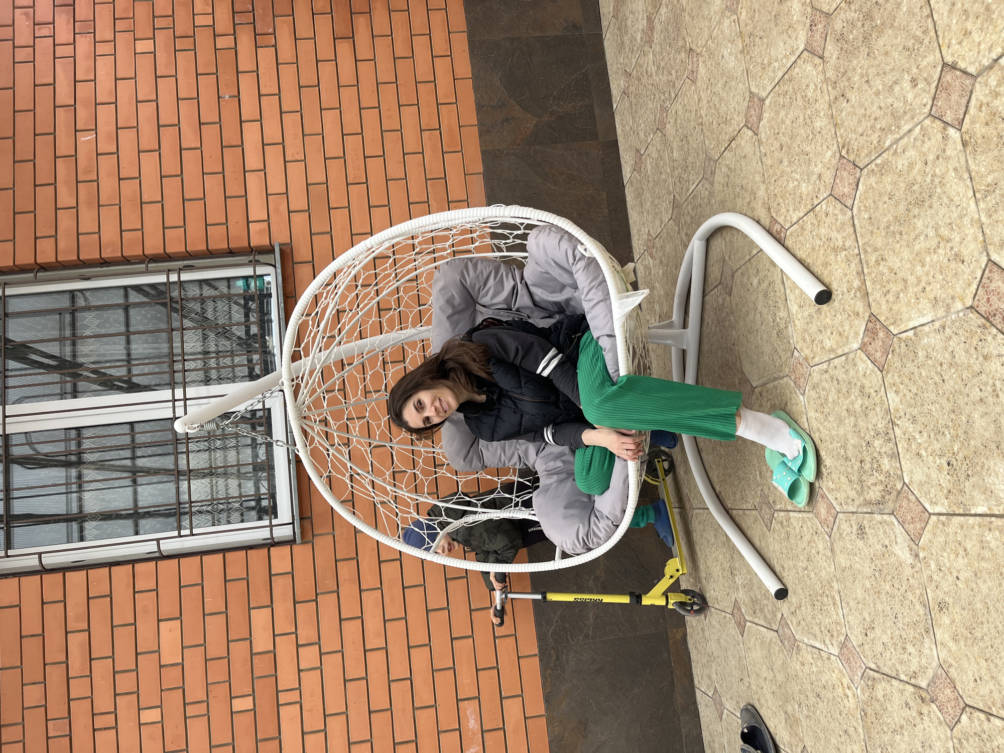 The image shows a woman and a young boy sitting in a white, egg-shaped swing chair located on a tiled patio next to a brick building with a barred window. The woman has long brown hair and is wearing a black jacket and green pants. The boy is sitting behind her in the chair and is wearing a blue hat and a dark jacket. A yellow scooter is positioned near the chair. The location is likely Kaspiysk, Russia, based on geolocation clues in the image metadata.