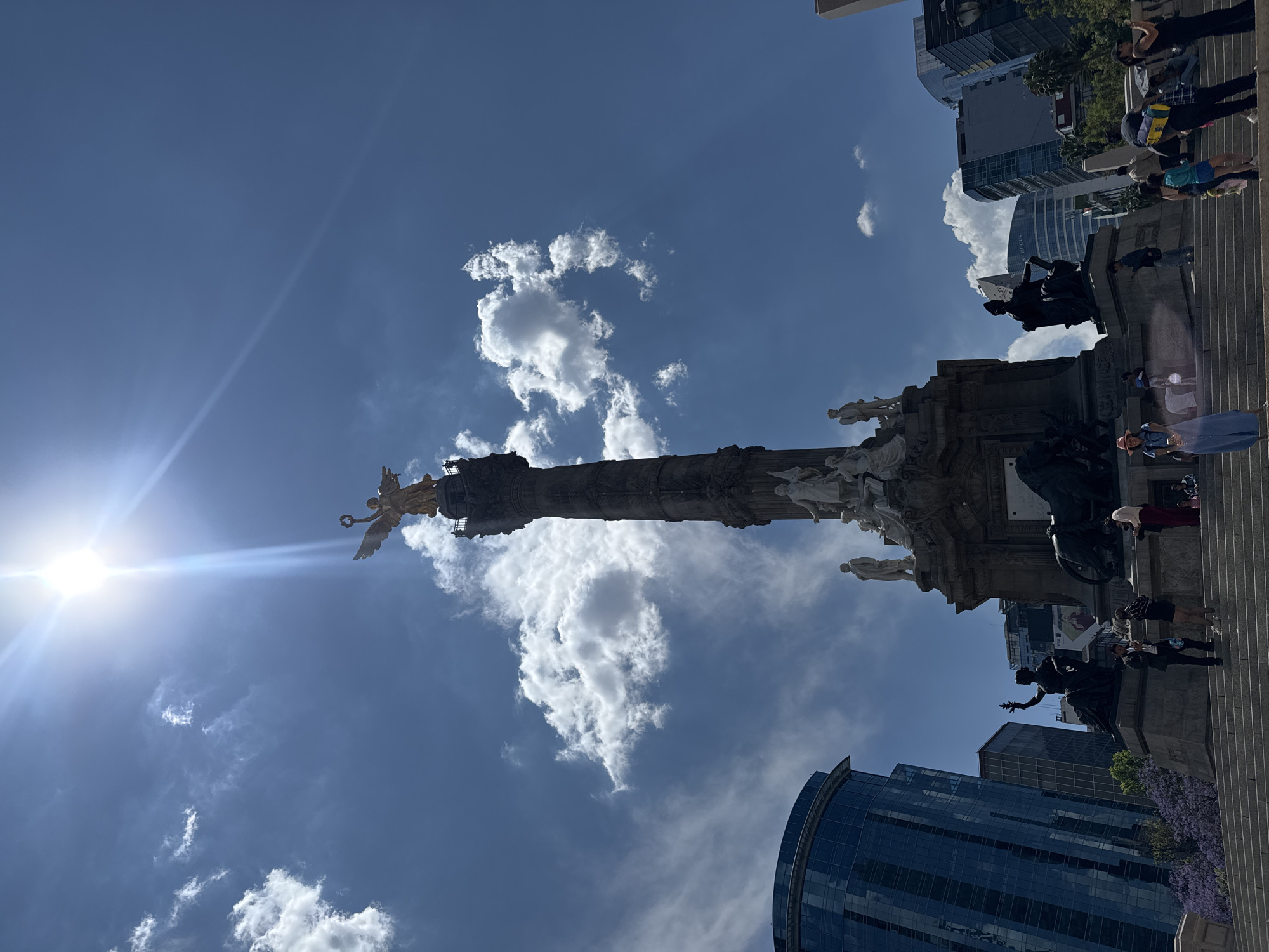 The image shows the Independence Monument in Mexico City, Mexico. The monument consists of a tall column topped with a golden statue of a winged Victory figure. At the base of the monument are several white statues, dark statues, and people. Some people appear to be climbing up to the monument's base. The sky is blue with scattered white clouds. The sun shines brightly, creating a lens flare effect. Modern buildings can be seen in the background.