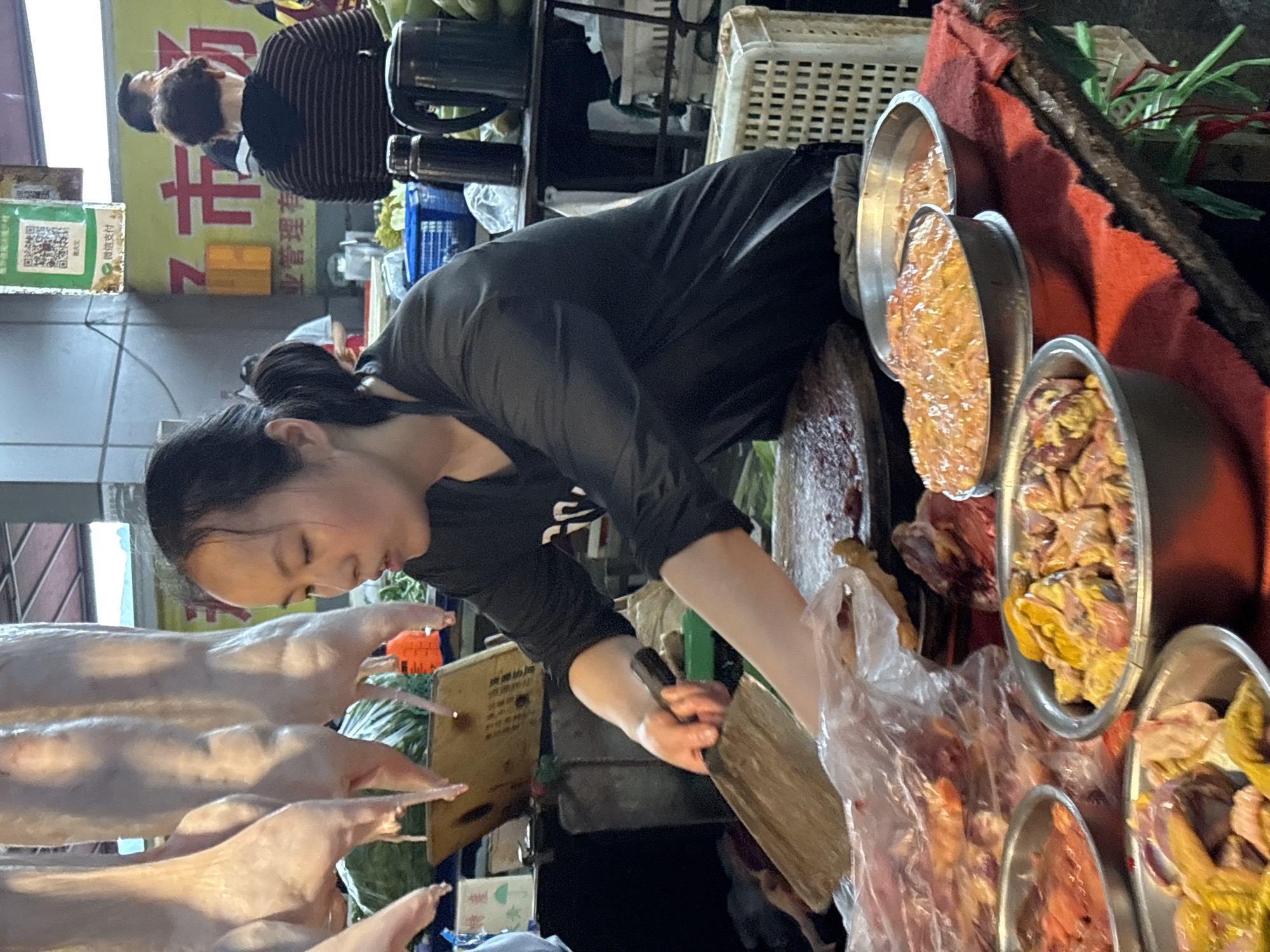 Here's a detailed description of the image:
**People:** A woman with dark hair pulled back in a bun is working at what appears to be a meat stall. She is wearing a dark long-sleeved shirt and is using a large knife and a wooden cutting board to handle the meat. Another person is visible in the background, wearing a striped shirt.
**Subjects/Objects:**
* Various cuts of raw meat are displayed in metal bowls. There are also several whole ducks (or similar poultry) laid out on a rack.
* A clear plastic bag is being used to package the meat.
* A knife and a wooden cutting board are being used.
* Other items such as baskets, bottles, and what appears to be a scale are visible in the background.
**Actions:** The woman is actively cutting and packaging meat for sale.
**Interactions:** The primary interaction is between the woman and the meat she is preparing. There's also an interaction between her and the customer.
**Environment:** The scene seems to be in an outdoor market. Based on the clues in the image, the location is the city of Jiangjin, China. The lighting is somewhat dim, suggesting the market is either indoors or it is evening. The overall impression is of a bustling, busy market environment.