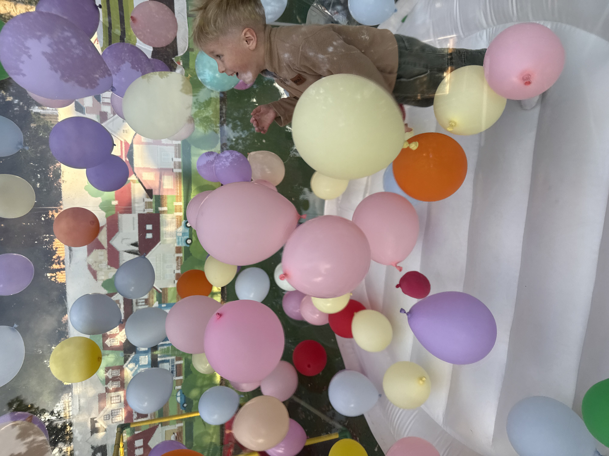 A young boy with blond hair, wearing a brown fleece shirt and green pants, is partially visible in the upper right quadrant of the image. He is looking to his left, with his mouth open as if speaking or laughing. The scene is filled with numerous colorful balloons of various sizes, including pastel purple, pink, yellow, orange, blue, and green. These balloons are floating in what appears to be an indoor setting, possibly a bounce house or a party area, given the presence of inflated white vertical structures in the background on the right.
The balloons are not evenly distributed, with some clustered together and others more spread out. The background, seen through the balloons and a reflective surface, appears to depict a somewhat blurred outdoor scene with buildings that resemble houses. The lighting suggests it is daytime. The overall atmosphere conveyed by the boy's expression and the vibrant balloons is one of fun and celebration. There is no visible text in the image.