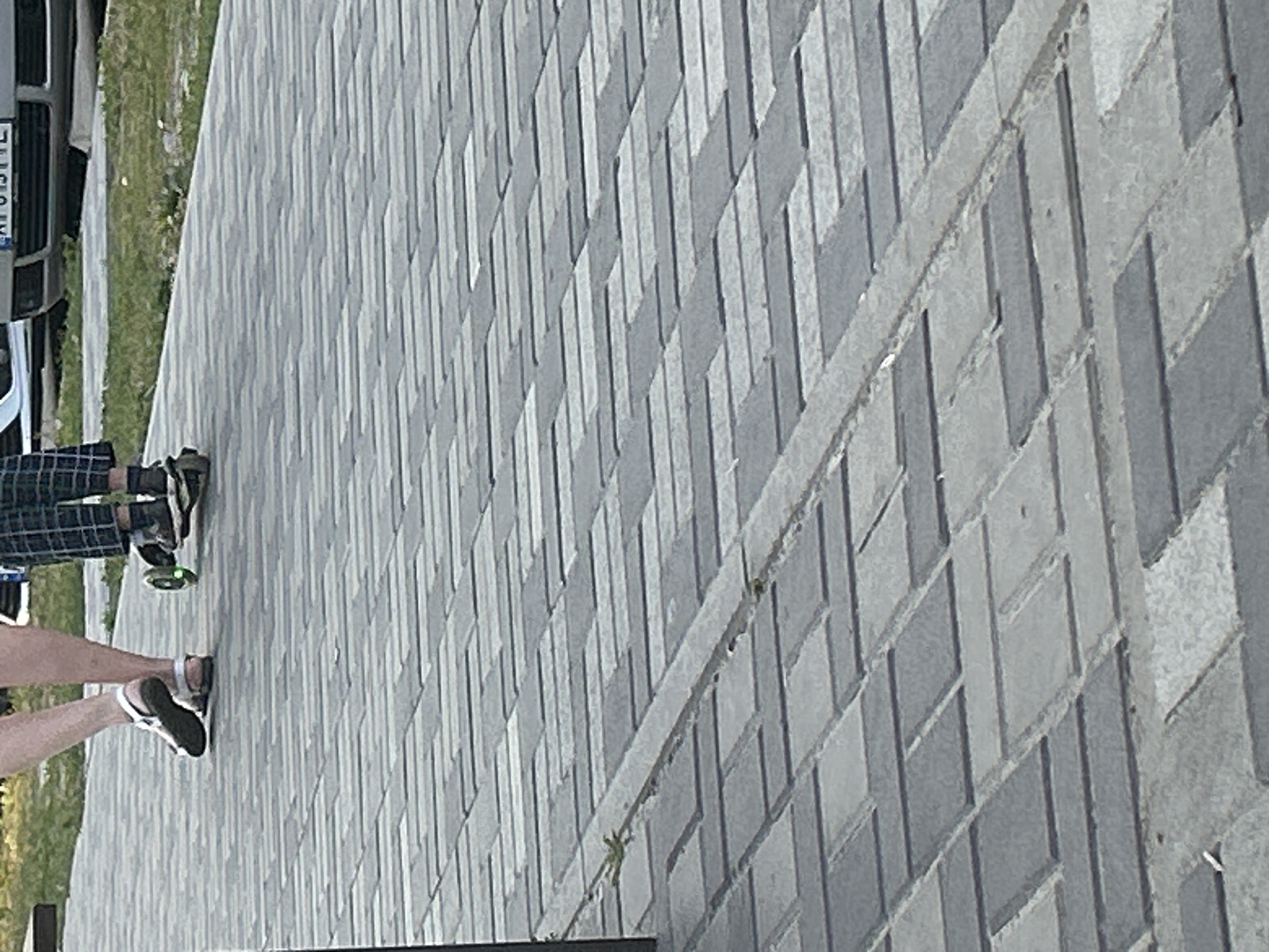 The image depicts a paved walkway with a distinct pattern of grey and darker grey rectangular pavers. In the upper left, two individuals are partially visible. The person on the left is wearing white sandals and has their legs and feet in the frame. To their right, another person is on a scooter, wearing checkered pants and dark socks. Their feet are on the scooter's deck. The background shows a grassy area and a portion of a car, including its license plate. The scene appears to be an outdoor public space, possibly a park or a street, in Brovary, Ukraine.