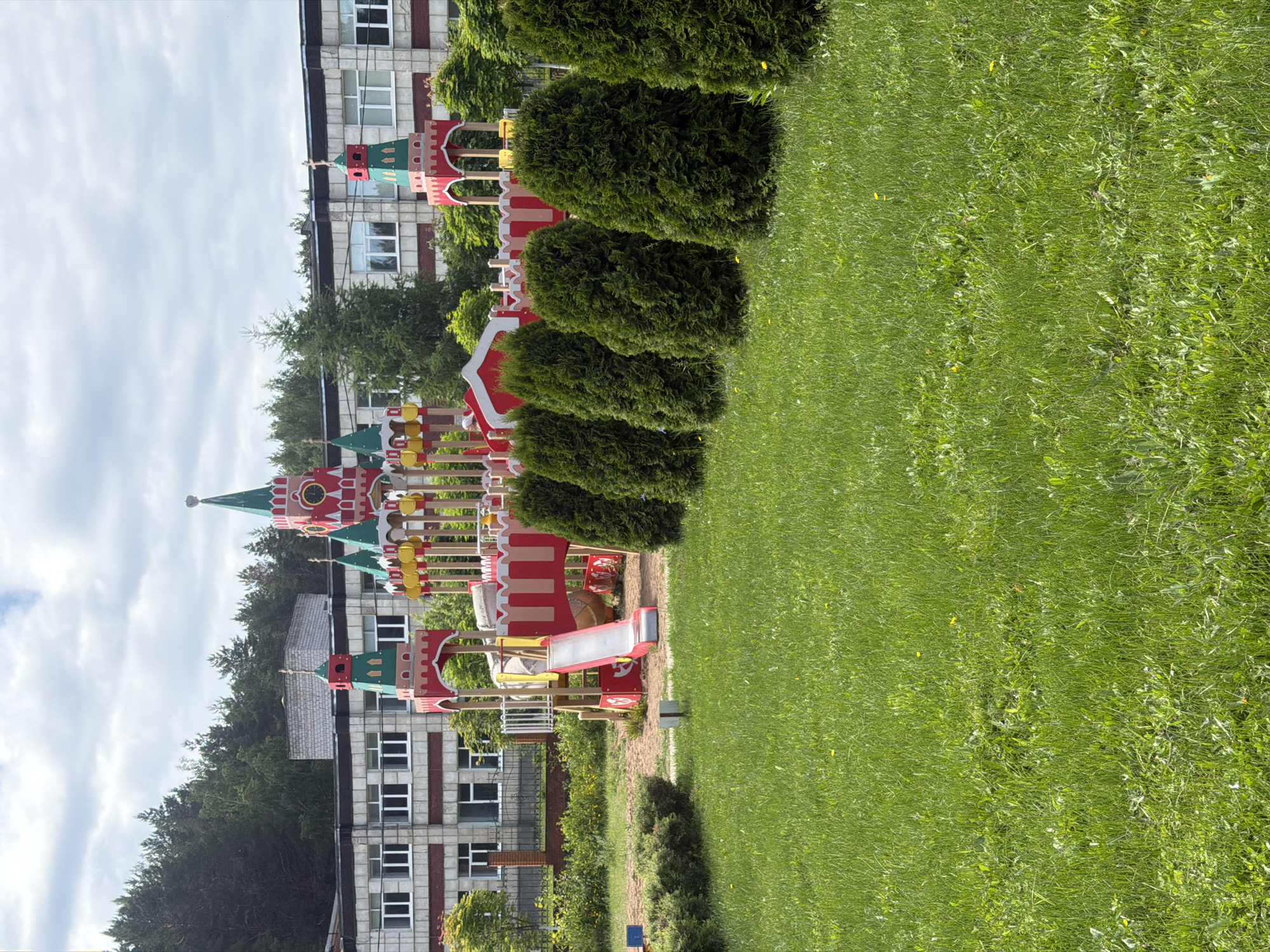 This image depicts a playground, designed to resemble the Moscow Kremlin, situated in front of a utilitarian building in the city of Chaikovsky, Russia. The scene is viewed under a partly cloudy sky.
The foreground is dominated by a bright green lawn, featuring varied grass lengths and interspersed dandelions. A row of five neatly trimmed, dark green conical evergreen shrubs extends across the mid-ground, forming a natural barrier between the lawn and the playground.
The playground itself is centrally located and constructed with a prominent red, green, and gold color scheme, mirroring Russian architectural styles. Notable features include a tall red tower with a green conical spire and a visible clock face on the right side of the structure. A smaller red and green tower, topped with a cross-like finial, is positioned closer to the center. A red slide with white railings descends from the main play structure into a sandy area on the left. No individuals are present on the playground.
In the background, a two-to-three-story building with a light-colored facade and multiple rectangular windows is visible behind a dark metal fence with brick posts. A dense forest of coniferous trees forms a dark green natural backdrop behind the building, extending into the upper left portion of the image. The sky above is overcast with a mix of white and grey clouds.