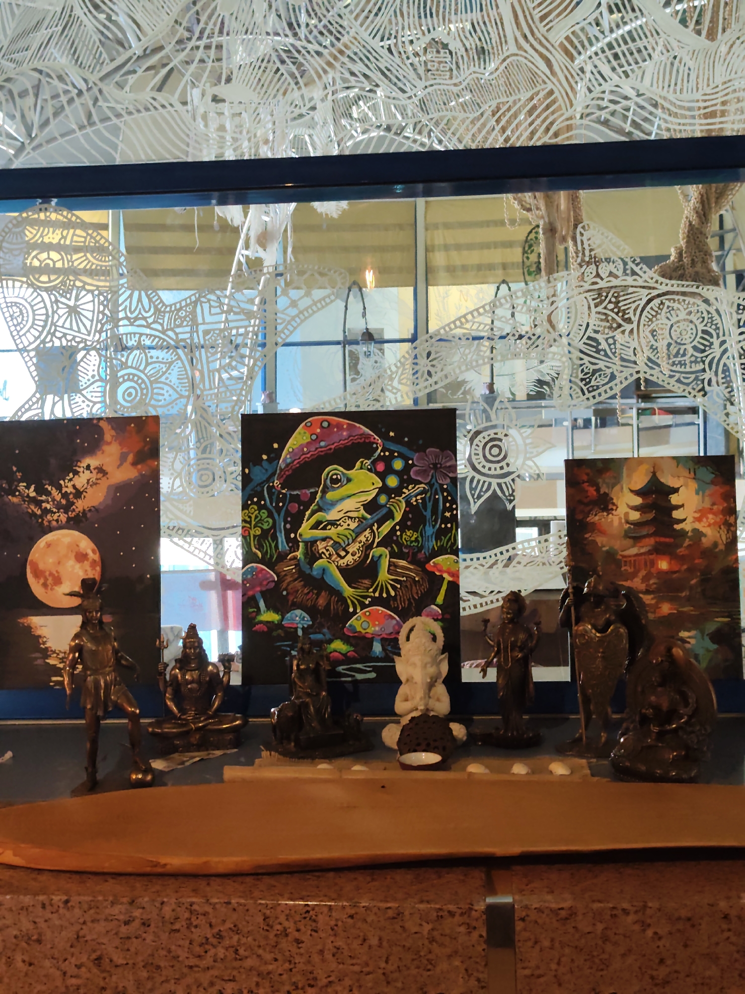 The image depicts a display of art and figurines on a shelf against a backdrop of ornate white cutouts and a window with a view of what appears to be an indoor public space with yellow blinds.
On the shelf, from left to right, are three paintings and several bronze and white figurines. The leftmost painting is a dark night scene with a large, vibrant orange moon and silhouetted trees. To its right is a painting of a green frog sitting under a colorful mushroom, playing a lute. The rightmost painting features a dark pagoda with fiery orange and red accents.
In front of the paintings are various figurines. The leftmost is a tall, slender bronze figure. Next to it is a seated bronze figure with multiple arms. Further right is a white figurine, possibly of a deity like Ganesha, seated next to a dark, round object with a small white bowl. There are two more bronze figurines to the right of the white one, one appearing to be armored and holding a shield, and another on the far right. The figurines are arranged on a flat, wooden surface that extends towards the viewer. The background suggests an indoor setting, possibly a cafe or shop. The white cutouts are intricate and decorative, resembling lace or elaborate patterns. The window behind the display offers a glimpse of a brightly lit area with yellow blinds, and further in the distance, what might be trees.
The scene is well-lit, highlighting the details of the artworks and figurines. There are no visible people in the frame. The overall impression is a curated display of decorative and artistic items.
This image was taken in Sochi, Russia, inside a venue identified as "Art-Cafe "Kalyug"".