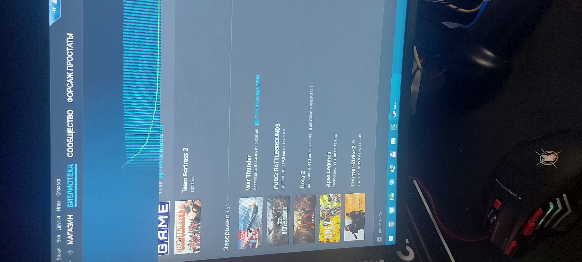The image shows a computer screen displaying the Steam client. The Steam client is in the "Library" section, showing a list of installed games. Some of the games listed are Team Fortress 2, War Thunder, PUBG: Battlegrounds, Dota 2, Apex Legends, and Counter-Strike 2. The download progress of these games is displayed. The upper part of the Steam client shows a download graph. The text is in Russian, indicating a Russian-speaking user. A computer mouse is also visible. The image provides no information about Tashkent, Uzbekistan.