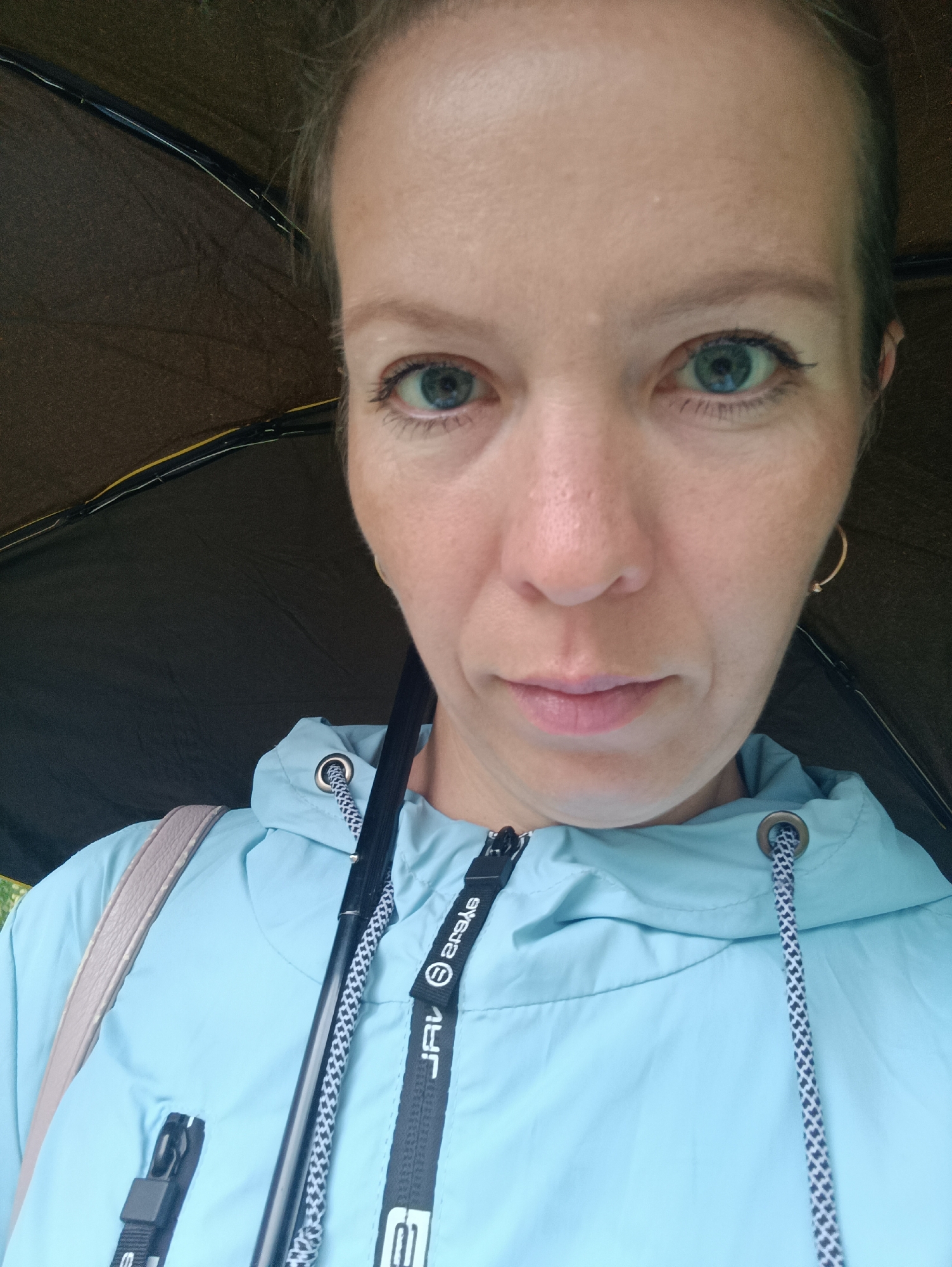 A close-up, eye-level selfie captures a woman under an open black umbrella in Chaikovsky, Russia. The woman, likely in her 30s or 40s, has fair skin, light-colored hair, and prominent light blue/green eyes, looking directly at the camera with a neutral expression. She is wearing a light blue hooded jacket with a black zipper.
Visible details of her attire include a black and white patterned drawstring on her jacket's hood, threaded through a metal eyelet. The black strap of her zipper pull shows the text "JRV" and "eyeS" (reversed). Another black strap, possibly from a lanyard or bag, is visible on the right side of her jacket with the text "aReke" (reversed). A light-colored shoulder strap, possibly from a purse or bag, is seen over her left shoulder.
The interior of the umbrella, positioned above her head, is dark brown/black with subtle yellow accents on its ribs, indicating its use due to likely overcast or rainy weather. The black stem or handle of the umbrella is central in the frame. The background is mostly obscured by the umbrella, with only a small hint of green visible on the far left. The overall lighting is soft and diffused.