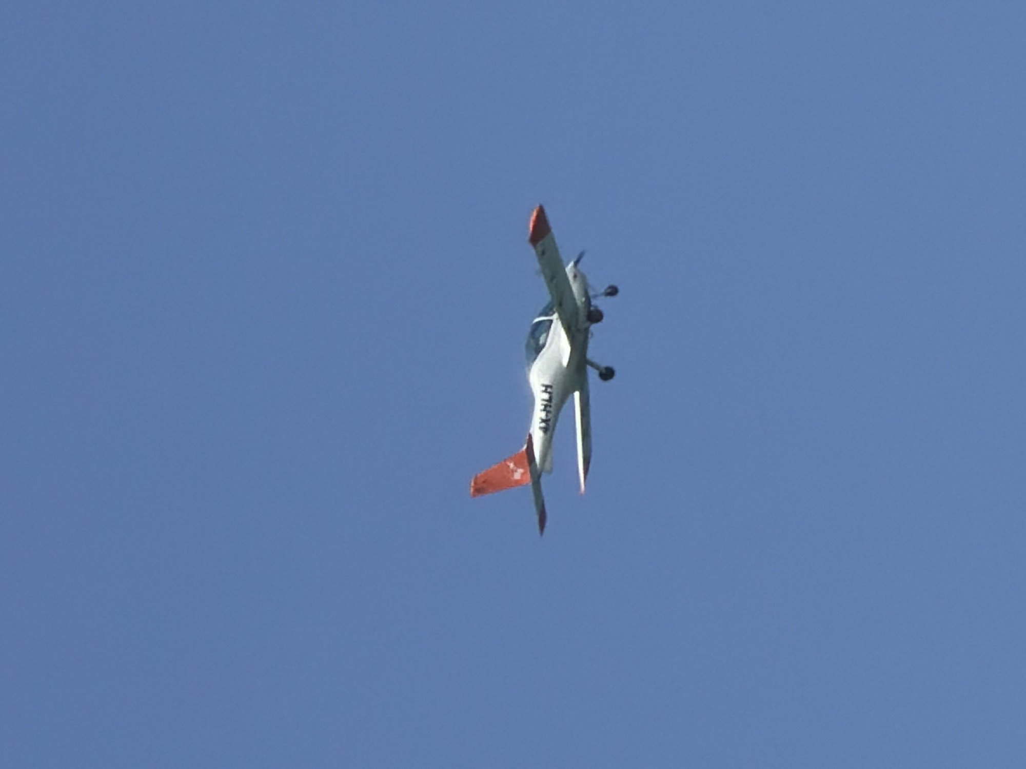 A white single-engine aircraft with red accents is flying in a clear blue sky. The registration number "4X-HLH" is visible on the tail of the plane. The image was likely taken outdoors during daylight. The location is not discernible, though the aircraft registration indicates it may be in Israel. There are no people visible in the image.