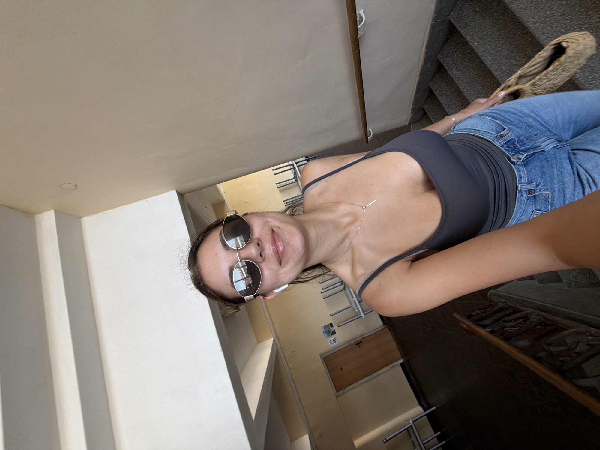 A young woman with sunglasses and a gray tank top stands on a staircase. She is holding a woven bag and wearing blue jeans. In the background, there is a hallway with doors and stacked chairs. The location is the staircase of a building in Burgas, Bulgaria.