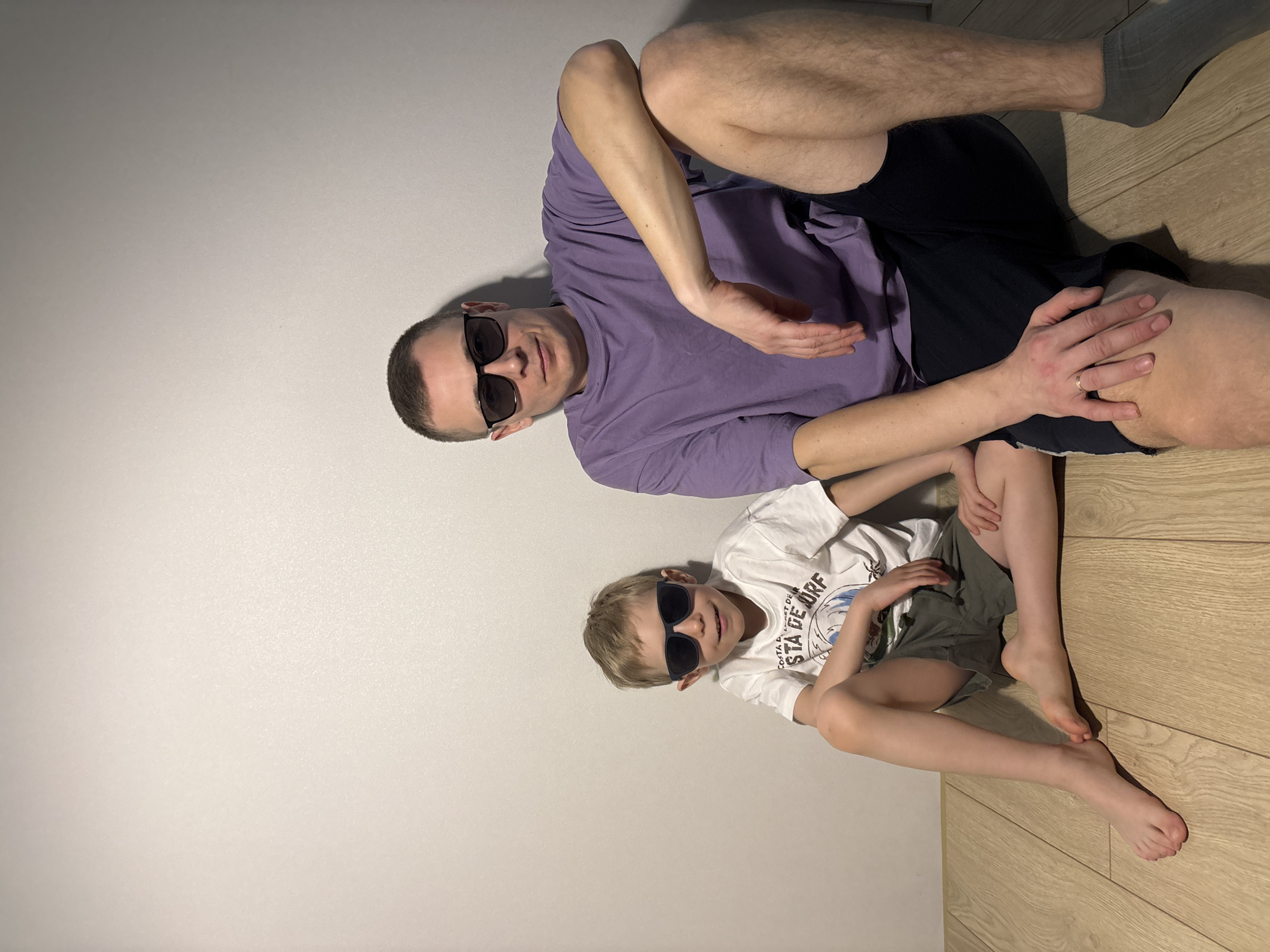 A rotated, high-angle indoor shot shows two males, an adult and a child, lying on a floor and against a wall. The image is oriented such that the subjects are lying vertically, with their heads towards the left side of the frame and feet towards the right. The setting appears to be a modern interior in Chaikovsky, Russia, characterized by a light grey or white wall and a light-colored wooden floor.
The adult, positioned higher in the frame, wears a purple short-sleeved t-shirt and dark shorts. He has short, dark hair and is wearing dark sunglasses, obscuring his eyes but showing a slight smile. His left arm is bent, hand resting near his chest, while his right arm extends towards the child. A gold wedding ring is visible on the ring finger of his right hand, which rests on the child's leg.
The child, positioned lower and partially embraced by the adult, wears a white short-sleeved t-shirt with dark graphics and dark shorts. Like the adult, the child wears dark sunglasses. Their light-colored hair is parted on the side, and a slight open-mouthed expression suggests a smile. Text visible on the child's t-shirt includes "COASTAL" and "STA DE ORF". Both subjects are barefoot, with their feet visible on the wooden floor. The overall mood appears relaxed and playful. The lighting is bright and even, casting soft shadows.