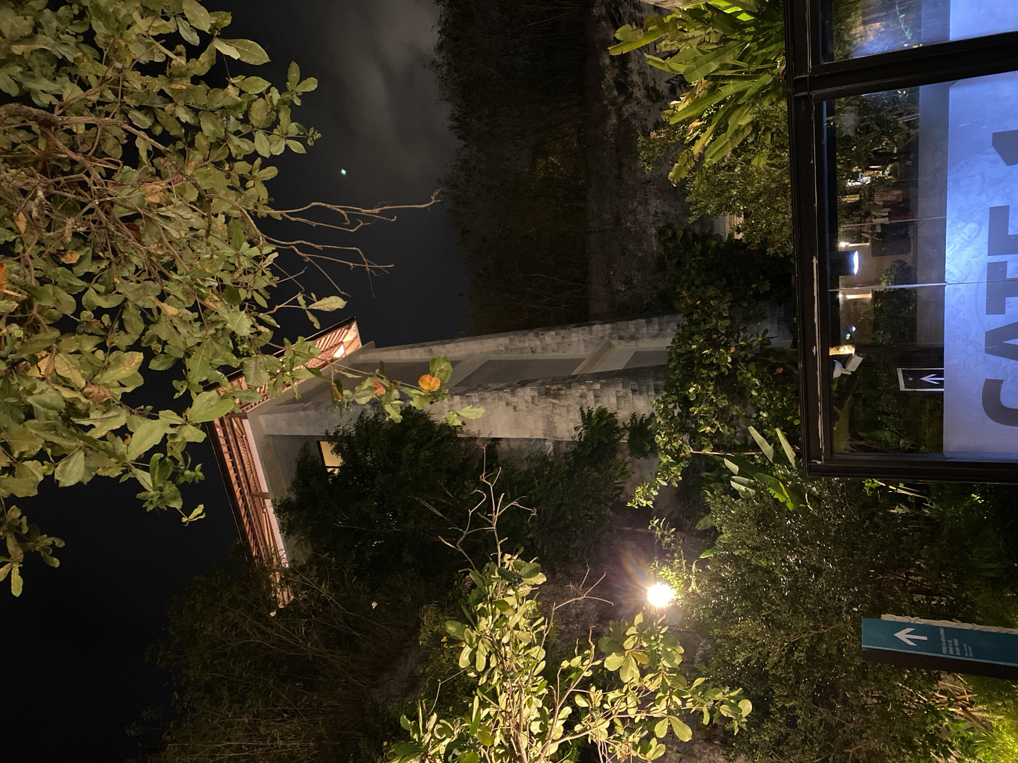 In the city of Kuta Selatan, Indonesia, at night, a tall, multi-story building with a tiled roof is visible amidst dense foliage and trees. The building's exterior appears to be made of textured concrete, with a few illuminated windows. A bright light source, possibly a streetlamp, illuminates some of the surrounding vegetation. In the foreground, a glass barrier with a sign that reads "GATE 1" is visible. Behind the barrier, indistinct figures can be seen, suggesting an indoor area. The overall scene is dimly lit, with the darkness of the night sky contrasting with the artificial lights.