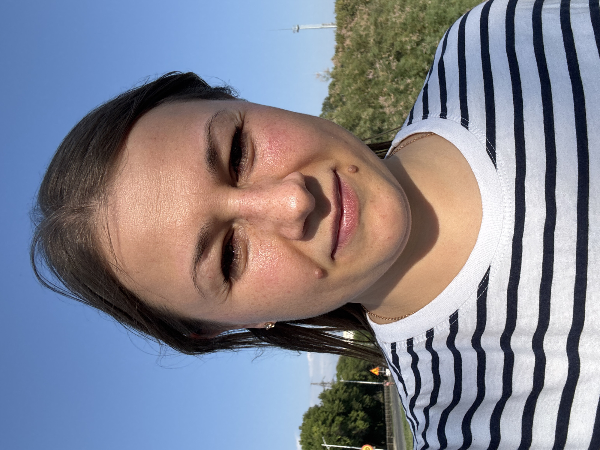 A woman is captured in a close-up selfie taken outdoors under a clear blue sky. She has dark hair pulled back, and her face is turned slightly towards the camera. Her skin shows a warm tone with visible freckles across her nose and cheeks, and some redness on her cheeks. She is wearing a striped t-shirt with dark blue and white horizontal stripes, and a delicate gold chain necklace is visible at her neckline. One side of her face is more prominently lit, highlighting features like her earlobe, which has a small, golden earring. In the background, to her left, a road with cars and traffic signs is visible, along with trees and utility poles. To her right, there is dense green foliage and a distant, tall, slender structure, possibly a radio tower or lighthouse. The overall impression is a candid portrait taken on a sunny day in an urban or suburban environment. The location is Yevpatoriya, Ukraine.