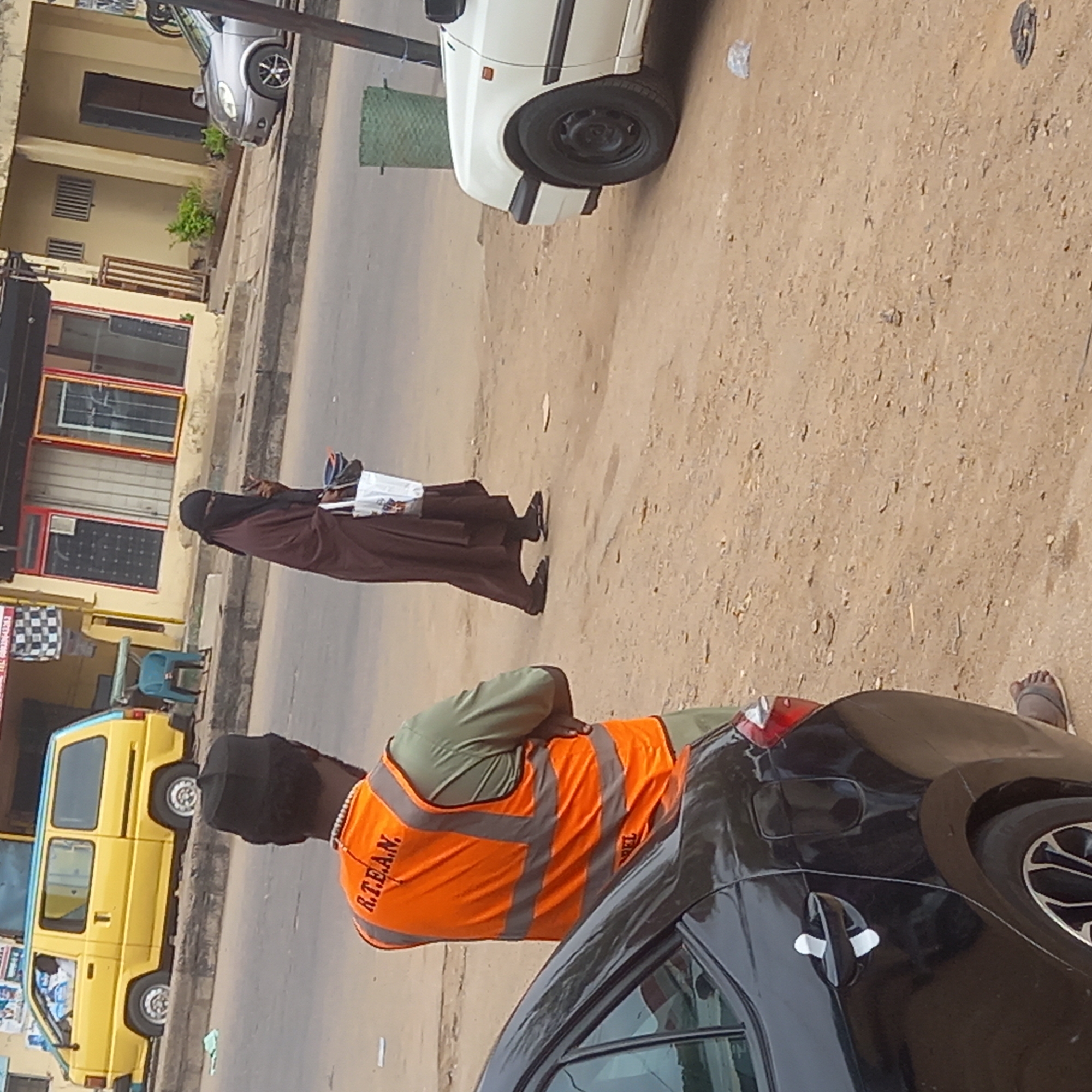 The scene is a street in Dadi Riba, Nigeria. A woman in a brown robe and niqab is walking. A man in an orange R.T.E.A.N. vest is standing near a black car. A yellow minibus is also present. Several cars are parked on the side of the road. Buildings line the street. The road surface is dusty.