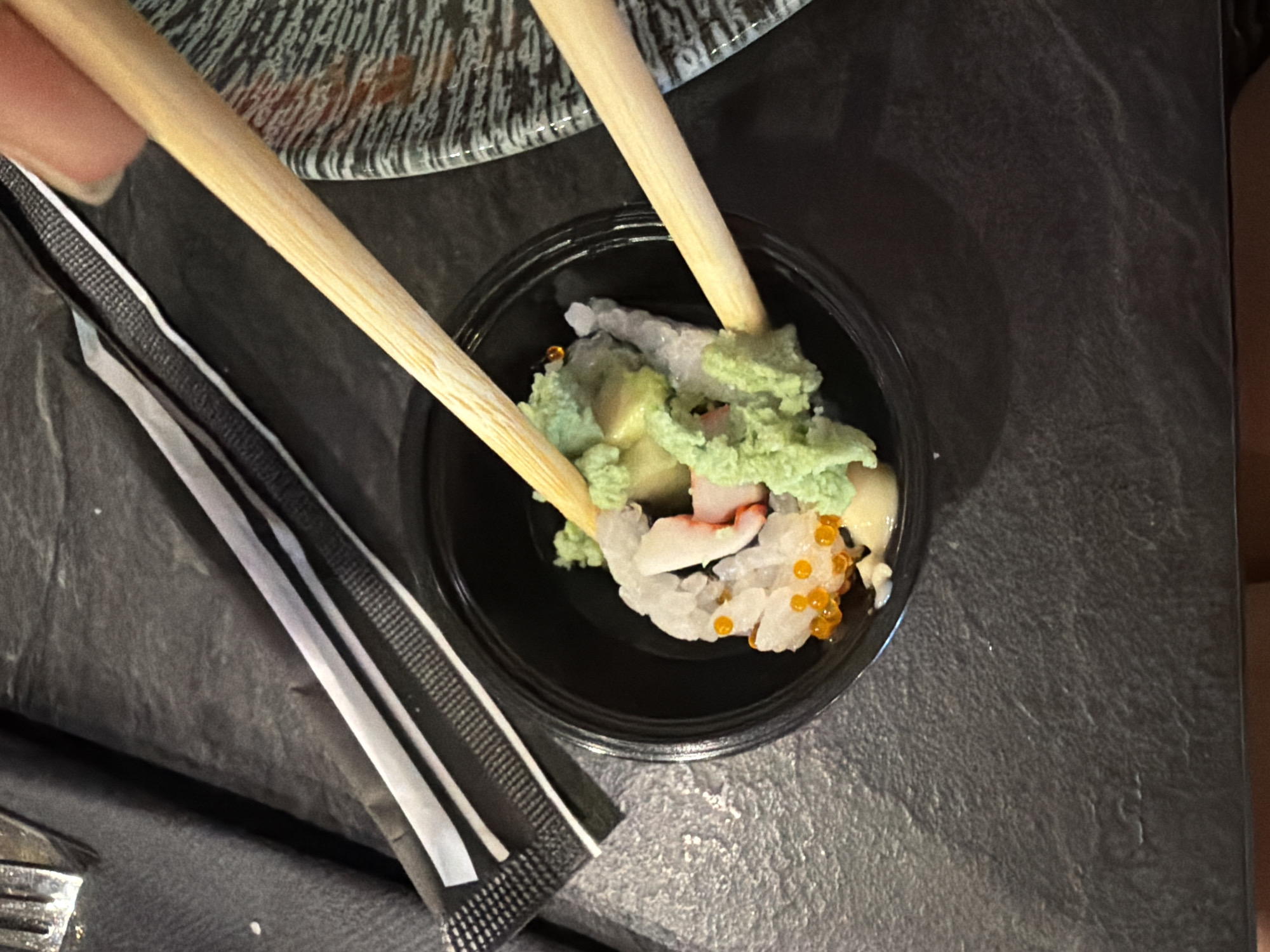 This image shows a close-up of a small black bowl containing sushi. The sushi appears to be a cut roll, with white rice, pinkish-white seafood, and bright orange roe. A dollop of bright green wasabi sits on top of the sushi. Two wooden chopsticks are being used to pick up a piece of the sushi. One chopstick is in the bowl, touching the sushi and wasabi, while the other is positioned above the bowl, also near the sushi. The bowl is resting on a dark, textured surface that appears to be a table. To the left of the bowl, a portion of a black and white wrapper is visible. To the right, a patterned plate with dark and light grey stripes is partially visible, with some reddish liquid staining it. The lighting suggests an indoor setting, possibly a restaurant. The image does not contain any information to place it in Kiti, Cyprus.