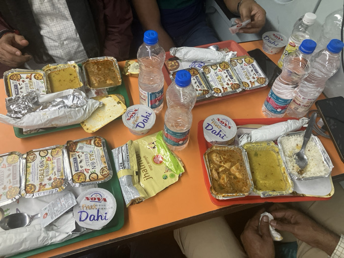 The image shows two green trays and one red tray filled with Indian meals and refreshments on an orange table. The meals consist of foil containers filled with curry, lentils, and rice, as well as flatbreads wrapped in foil. There are several water bottles and tubs of yogurt labeled "Dahi". Some of the food containers are labeled "MEALS ON WHEELS." Also visible are packets of "jhalmuri" snack. The scene appears to be a train journey where passengers are enjoying a meal.