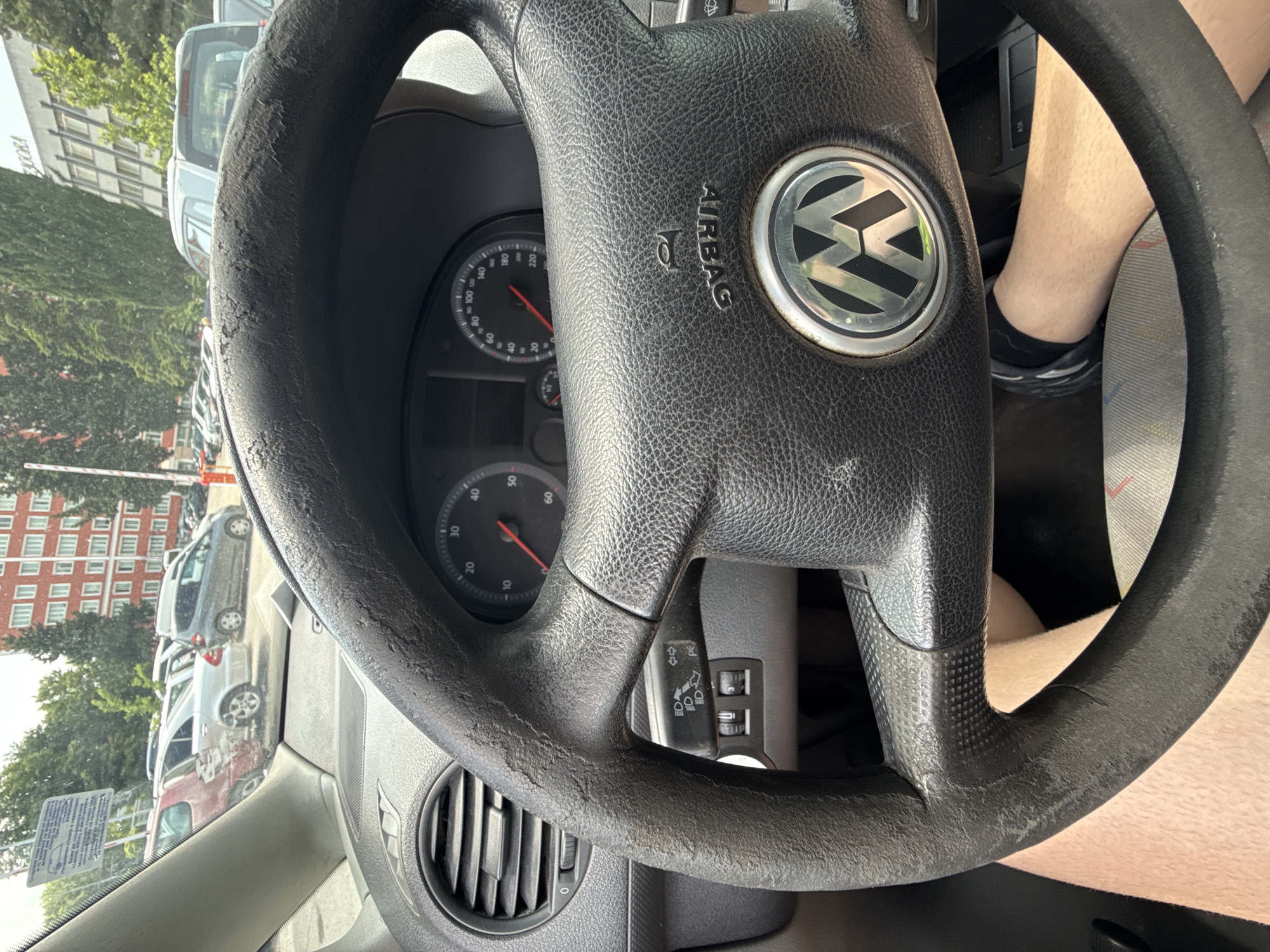 The image shows the interior of a car from the driver's perspective, looking out towards an urban scene in Stara Zagora, Bulgaria. The primary subject is a black Volkswagen steering wheel, prominent in the foreground, which exhibits considerable wear and peeling on its surface, particularly around the rim. The Volkswagen logo is clearly visible in the center, along with the embossed word "AIRBAG" above it.
Behind the steering wheel, parts of the dashboard and instrument cluster are visible, including a speedometer with a red needle resting near zero, indicating the car is stationary. An air vent is located to the left of the steering wheel, and a stalk for lights or wipers is seen to the right. The driver's lower legs, wearing dark socks and shoes, are partially visible in the bottom right corner.
Beyond the windshield, the scene is bright, suggesting daytime with clear weather. Numerous vehicles, including cars and possibly vans, are parked in rows along what appears to be a street or parking lot. Green trees line the background, and several buildings are visible. One building features a sign with the word "ANCORA." Another building is characterized by red brick architecture. In the lower left, a white barrier and a sign with largely illegible Cyrillic text are present. The OCR analysis of this sign yields fragmented words such as "ED," "EDP," and "JOOKA." An "ASR" symbol is also visible near a button on the car's dashboard.