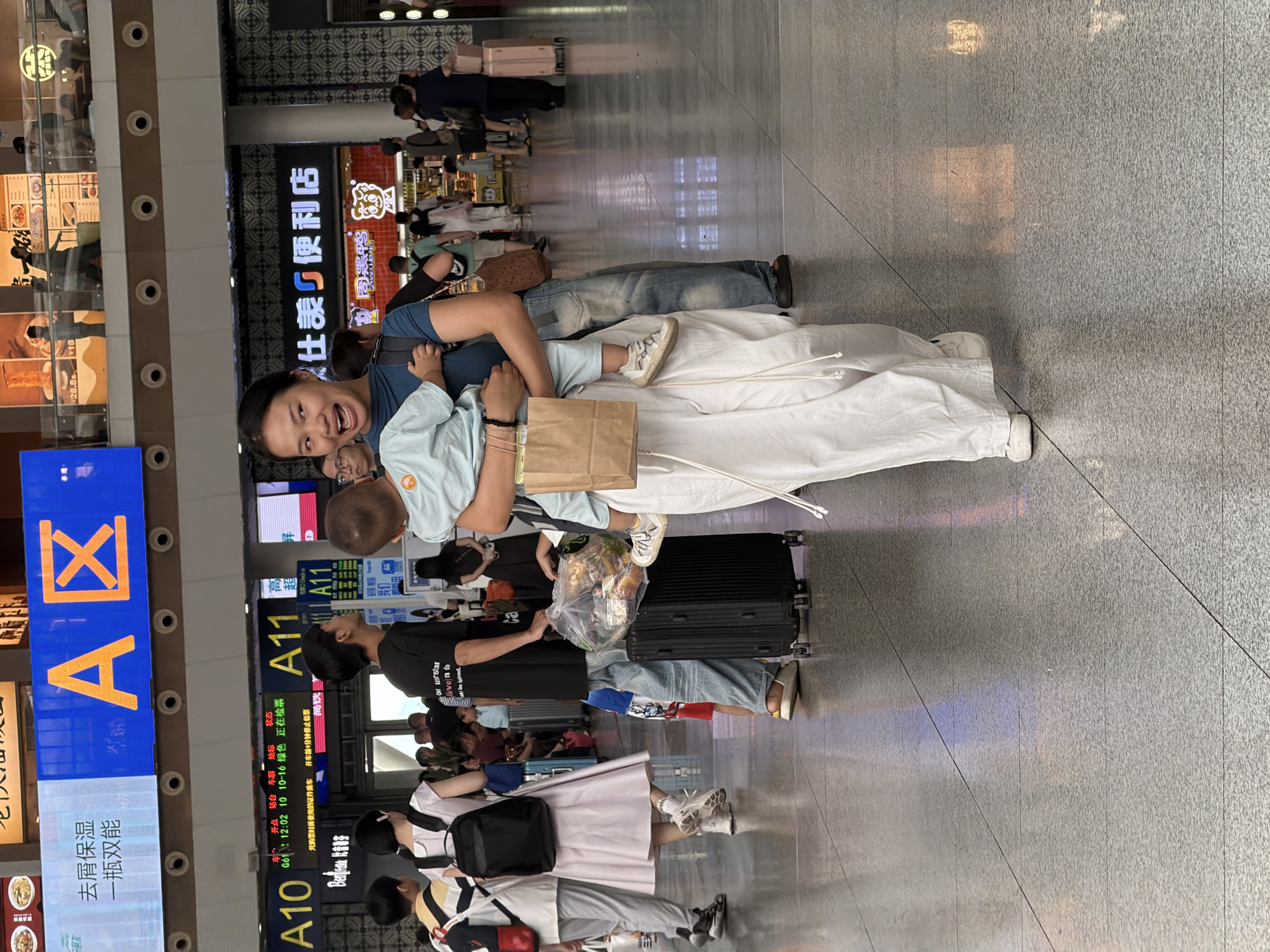 In Panyu District, China, a woman is holding a baby while walking through a busy indoor terminal, possibly a train station or airport, indicated by the large signs displaying "A10" and "A11" and the presence of luggage. The woman is wearing a dark blue t-shirt and loose white pants, and the baby is dressed in a light blue outfit. The environment is brightly lit with a polished floor reflecting the overhead lights and displays. Other individuals are visible in the background, some with luggage, suggesting a travel hub. The woman appears to be speaking or reacting to something with an open mouth and engaged expression.