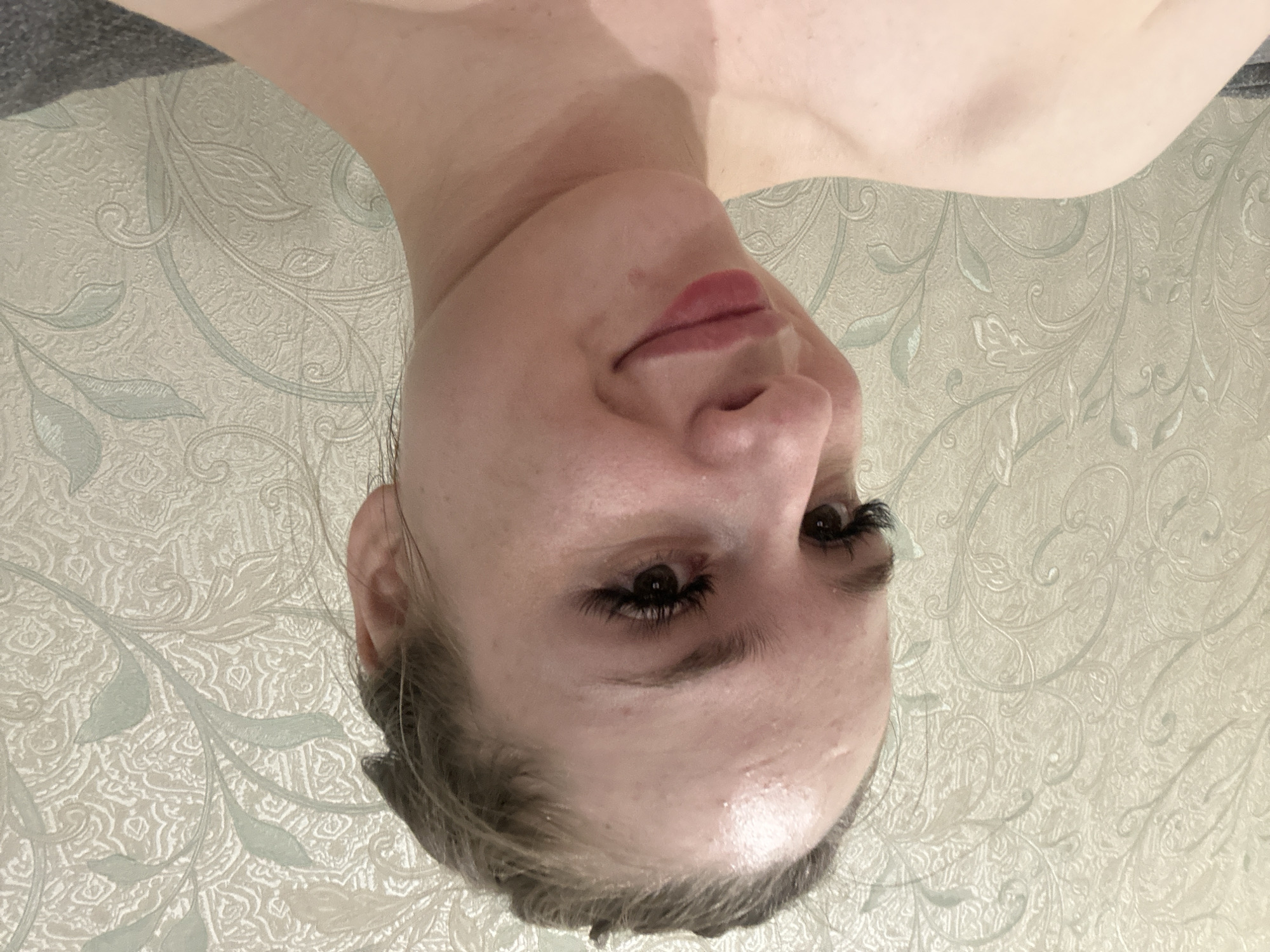 A young Caucasian woman with blonde hair pulled back is pictured in a close-up, head-and-shoulders shot. She is looking towards the viewer's left and smiling faintly. Her skin appears slightly oily on her forehead, and she is wearing false eyelashes and lip gloss. The background is a light-colored wallpaper with a scrolling floral pattern in muted greens and beige. The lighting is soft, and the image is slightly blurry.