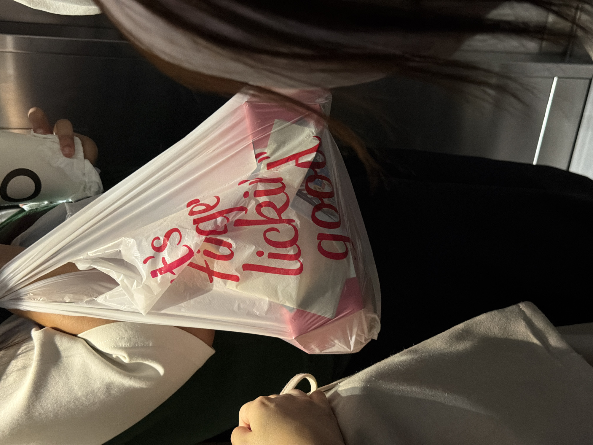 The image displays a close-up, angled perspective within a dimly lit indoor or vehicle setting. The central subject is a translucent plastic shopping bag, held loosely or resting. Prominent red text on the bag reads "it's finger lickin good". Inside the bag, various items in white and pink are discernible, likely packaging or products.
Parts of a person are visible in the frame. On the left side, a hand holds a soft, white object featuring a black circular design. Another hand is seen on the bottom right, gently grasping a loop of light-colored fabric, possibly from a blanket or piece of clothing. The individual appears to be wearing a light-colored, possibly off-white, top. Dark hair extends across the top of the frame.
The background is predominantly dark and indistinct, suggesting an interior space such as a car, bus, or train cabin, or a room with low illumination. The lack of natural light indicates it is likely nighttime or a heavily shaded indoor environment. There are no specific visual cues within the image itself to identify Chiang Mai City Municipality, Thailand, beyond the provided context. The scene captures a quiet moment, possibly during transit or rest, with personal belongings.