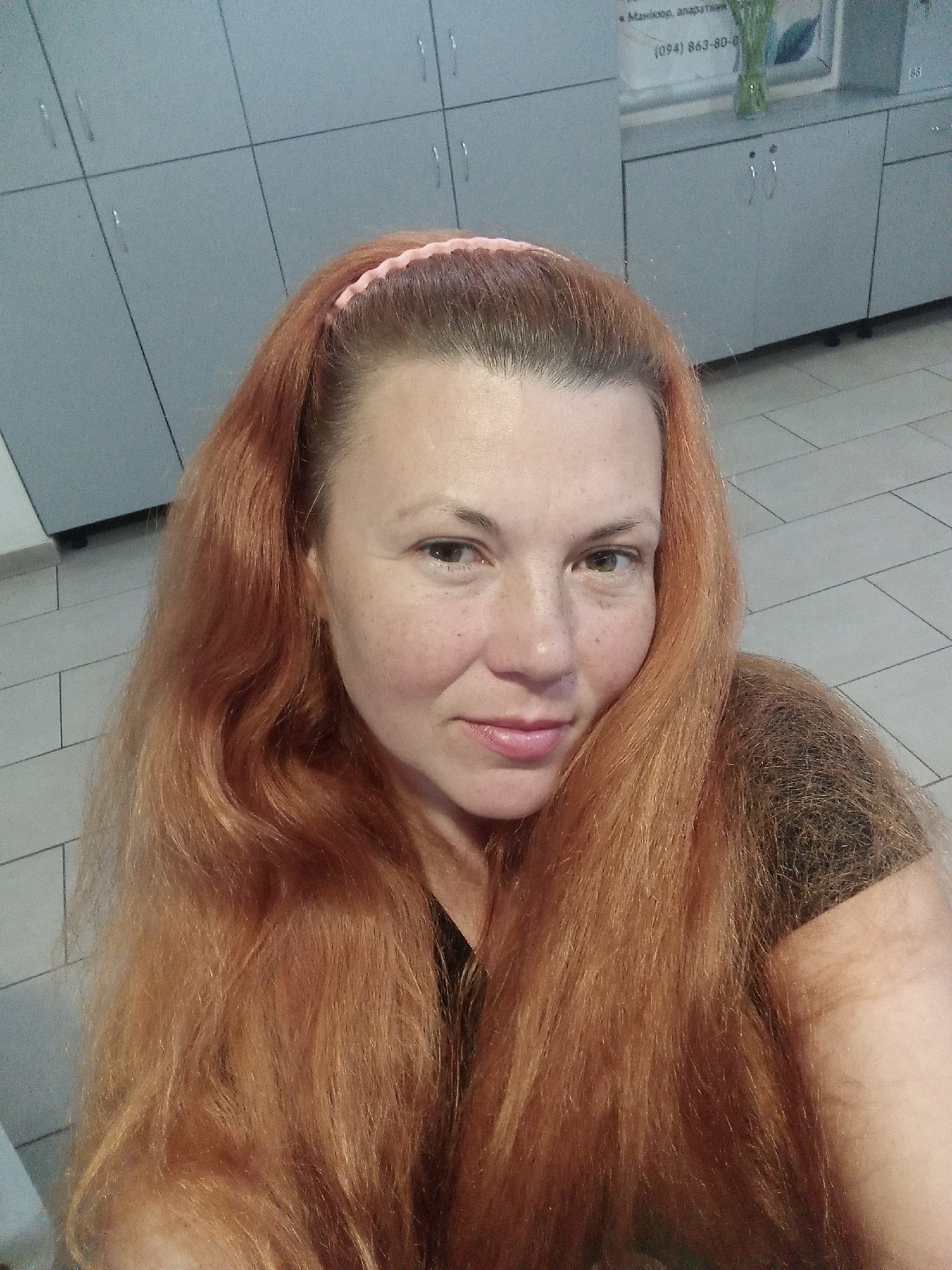 A medium shot shows a white woman with long, wavy auburn hair and fair skin, a light dusting of freckles across her nose and cheeks, and rosy lips, wearing a pink headband. She is looking directly at the camera with a slight smile. The background consists of light gray lockers and a tiled floor, suggesting an indoor setting such as a gym or spa. A partial view of text and a phone number on a poster is visible on the wall behind her. The lighting is artificial and casts a soft glow.