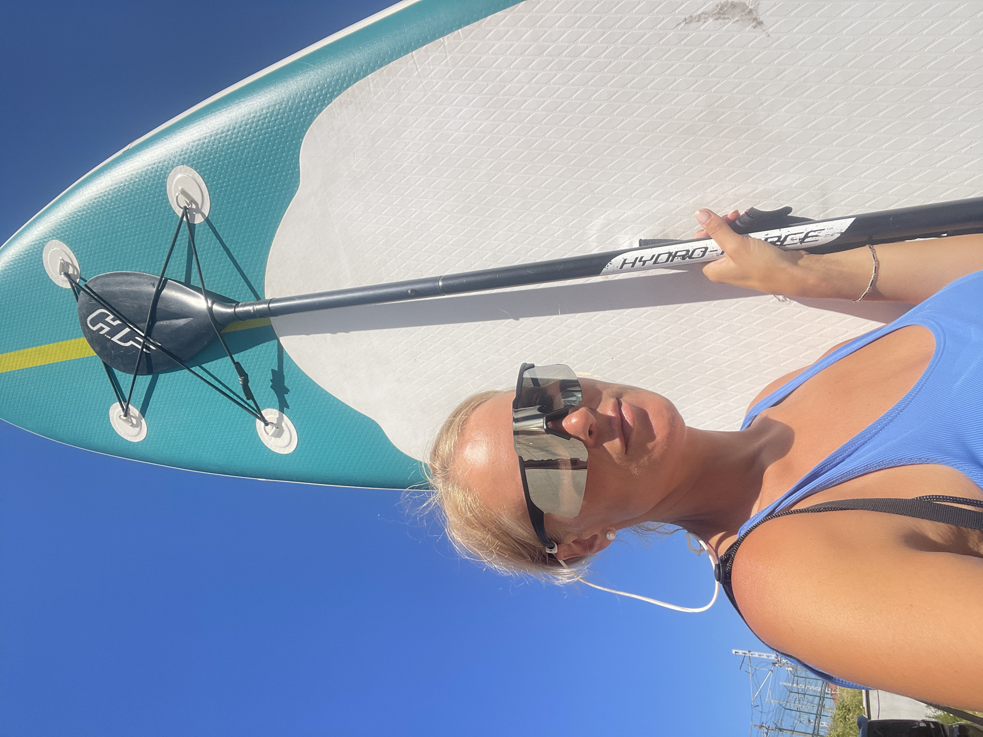 A blonde woman stands outdoors under a clear blue sky, holding a paddleboard. She is wearing oversized reflective sunglasses, a pearl earring, and a blue tank top. The paddleboard is teal and white with a black paddle attached. The scene appears to be in Sochi, Russia, as indicated by the presence of overhead power lines and a railway track in the background. The lighting suggests it is daytime.