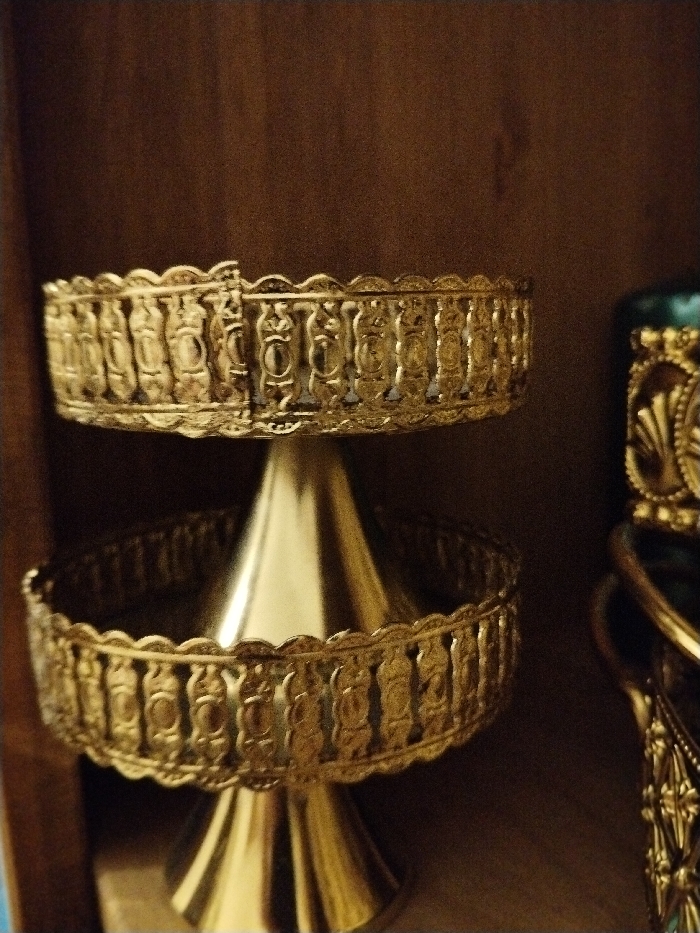A two-tiered golden metal decorative stand features ornate detailing around the edges of each tier. The stand has a cone-shaped stem. Part of a second golden object, also ornate, is partially visible on the right side of the frame. The objects are placed against a solid brown surface that is out of focus.