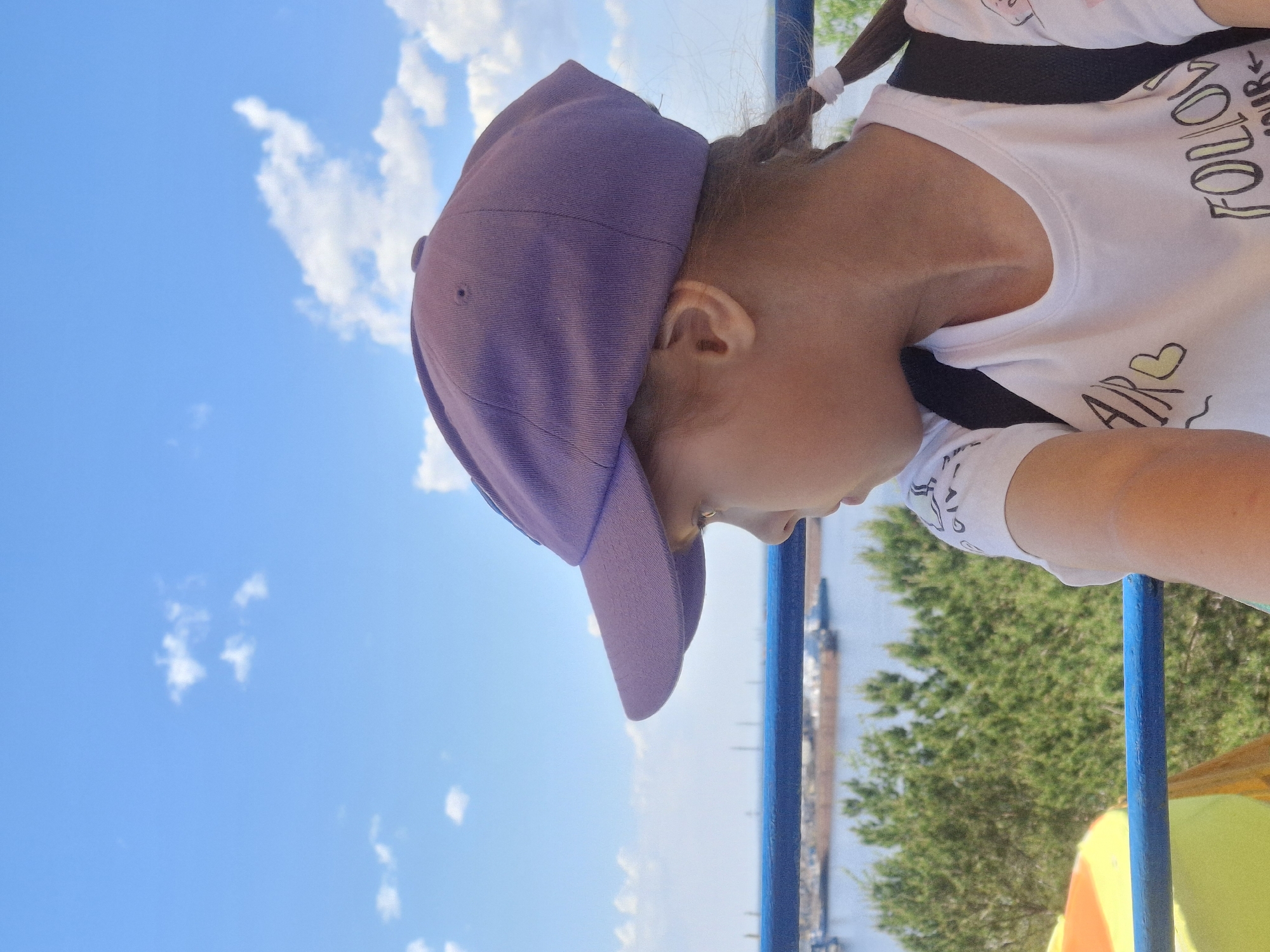 A young girl wearing a purple baseball cap and a white t-shirt with "FOLLOW YOUR" printed on it stands outdoors, likely near the city of Chaikovsky, Russia, based on the background scenery. The girl has a braid with white hair ties. She appears to be looking down, possibly leaning over a blue railing. Beyond the railing, there is a view of a river or reservoir, a building resembling a factory or industrial structure, and greenery including trees. The sky is blue with scattered white clouds.