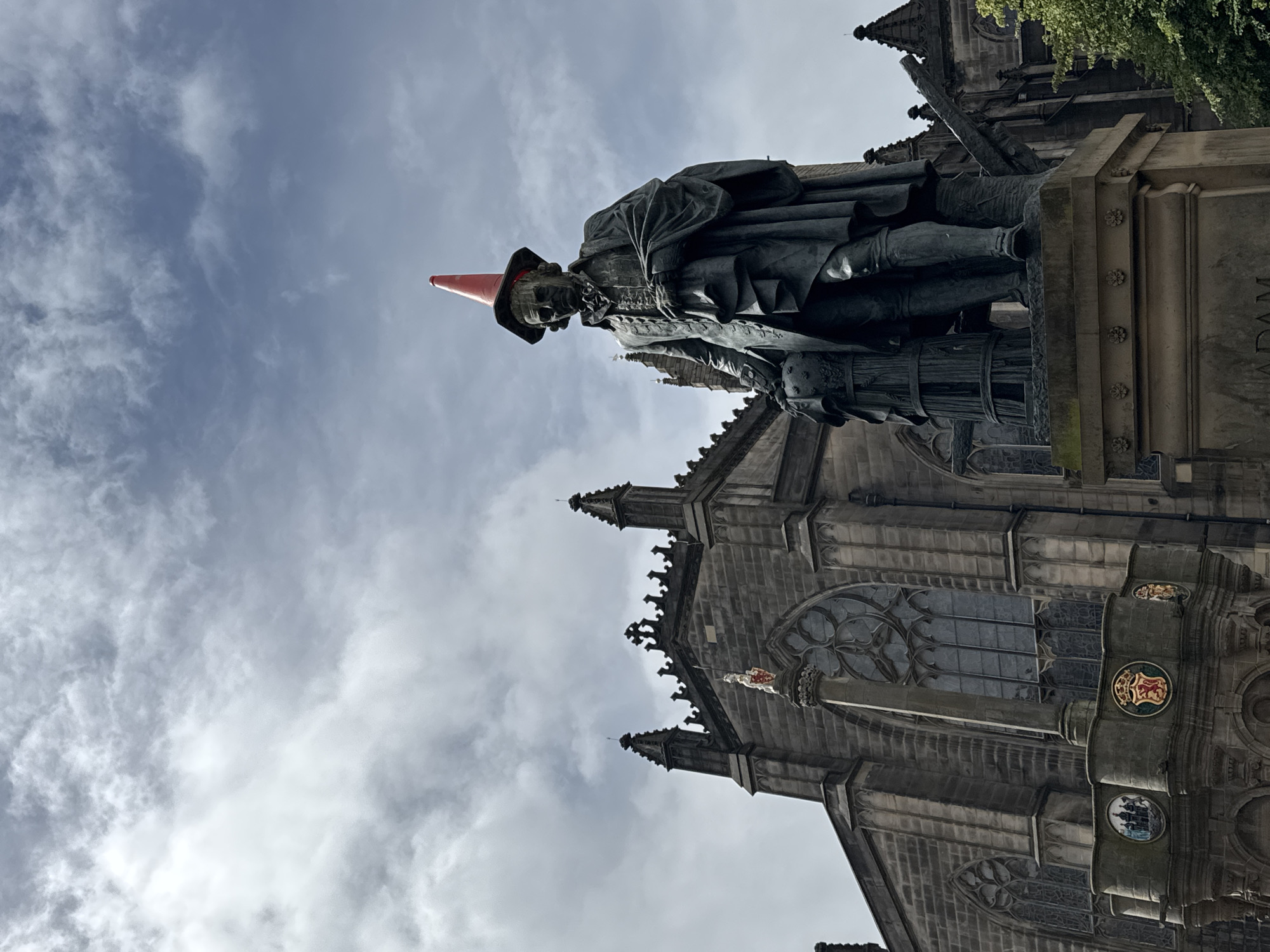 The photograph captures a bronze statue of Adam Smith, a prominent Scottish economist and philosopher, situated in the City of Edinburgh, United Kingdom. The statue is prominently positioned in the foreground, with a large Gothic-style building, likely a church or cathedral, serving as the backdrop. A striking detail is a bright orange traffic cone placed on top of the statue's head, suggesting a playful act of vandalism or a local tradition. The sky above is partly cloudy, with a mix of grey and white clouds against a blue sky. The statue is detailed, showing Smith in period attire, holding what appears to be a globe and a scroll. The building behind is made of stone and features architectural elements such as arched windows and spires. The overall lighting suggests it is daytime, possibly overcast.