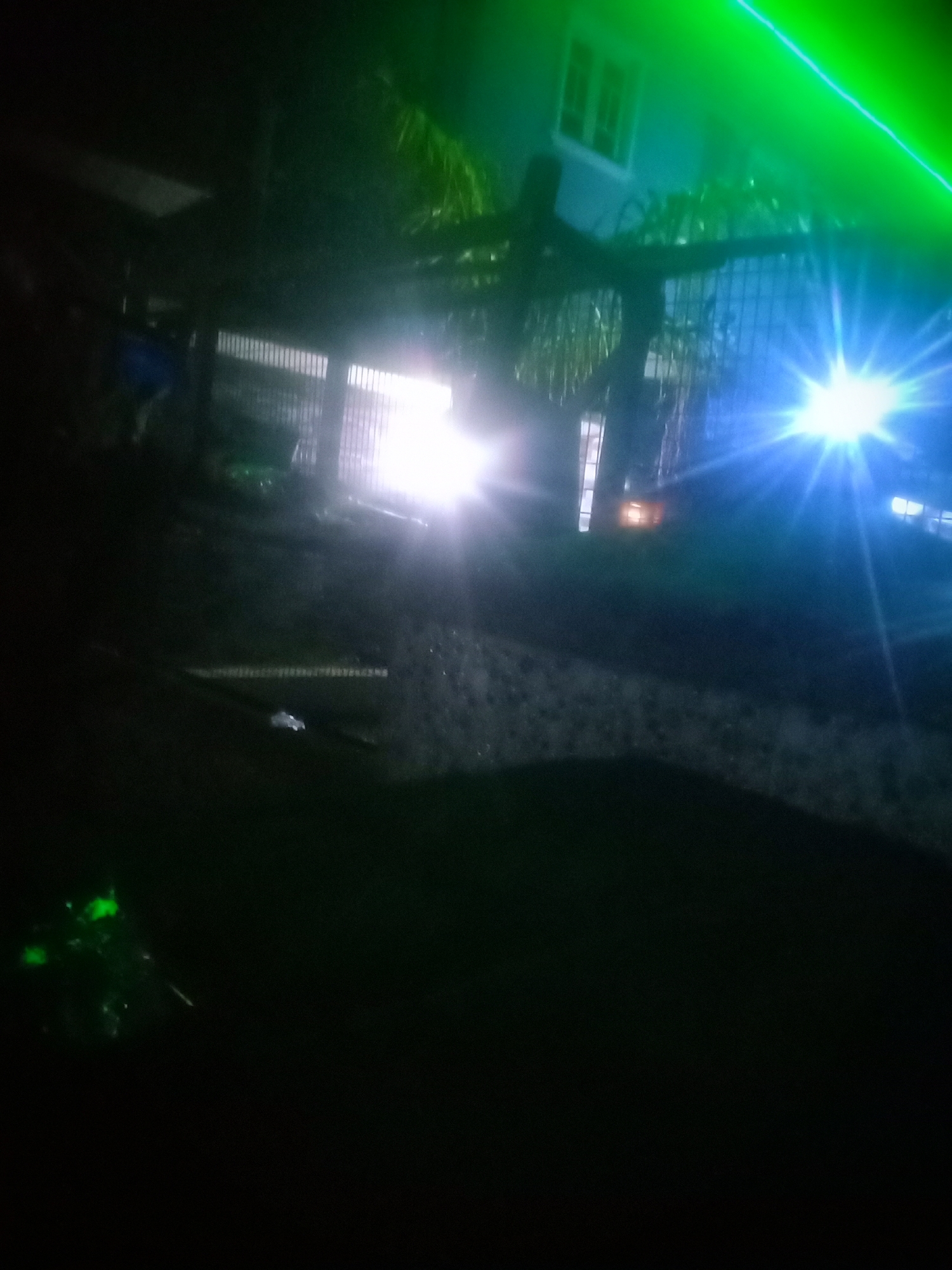The image captures a nighttime scene in an outdoor urban setting, likely a street or driveway. The foreground is dominated by darkness, with a faint green glow emanating from the lower left corner, suggesting an object with phosphorescent properties. In the middle ground, a bright white light, possibly from headlights or a streetlamp, creates a starburst effect, illuminating a section of the ground and a fence. Beyond the fence, a building with illuminated windows and a darker facade is visible, partially obscured by foliage. A green light beam streaks across the top of the image, originating from the upper right. The overall impression is one of low visibility with stark contrasts between illuminated areas and deep shadows.