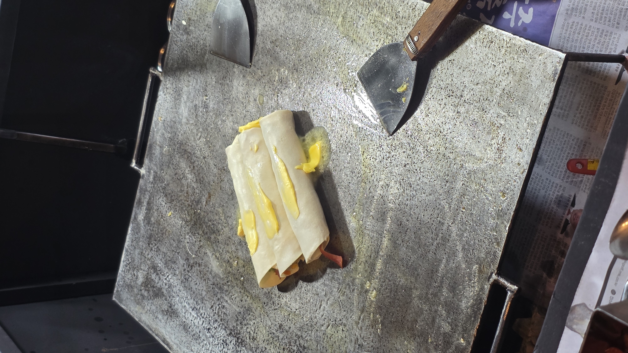 The image shows a metal griddle, likely part of a food cart or vendor setup. On the griddle are three rolled food items. They appear to be crepes, tortillas, or similar wrappers, filled with what appears to be ham and cheese. There are what appears to be dollops of cheese sauce on top. Two metal spatulas sit nearby on the griddle. To the right, there appears to be a section of newspaper. The food items are being prepared or cooked. The environment is a food service setting. The precise location within the city of Palohigeuh, Indonesia cannot be determined with the given information.