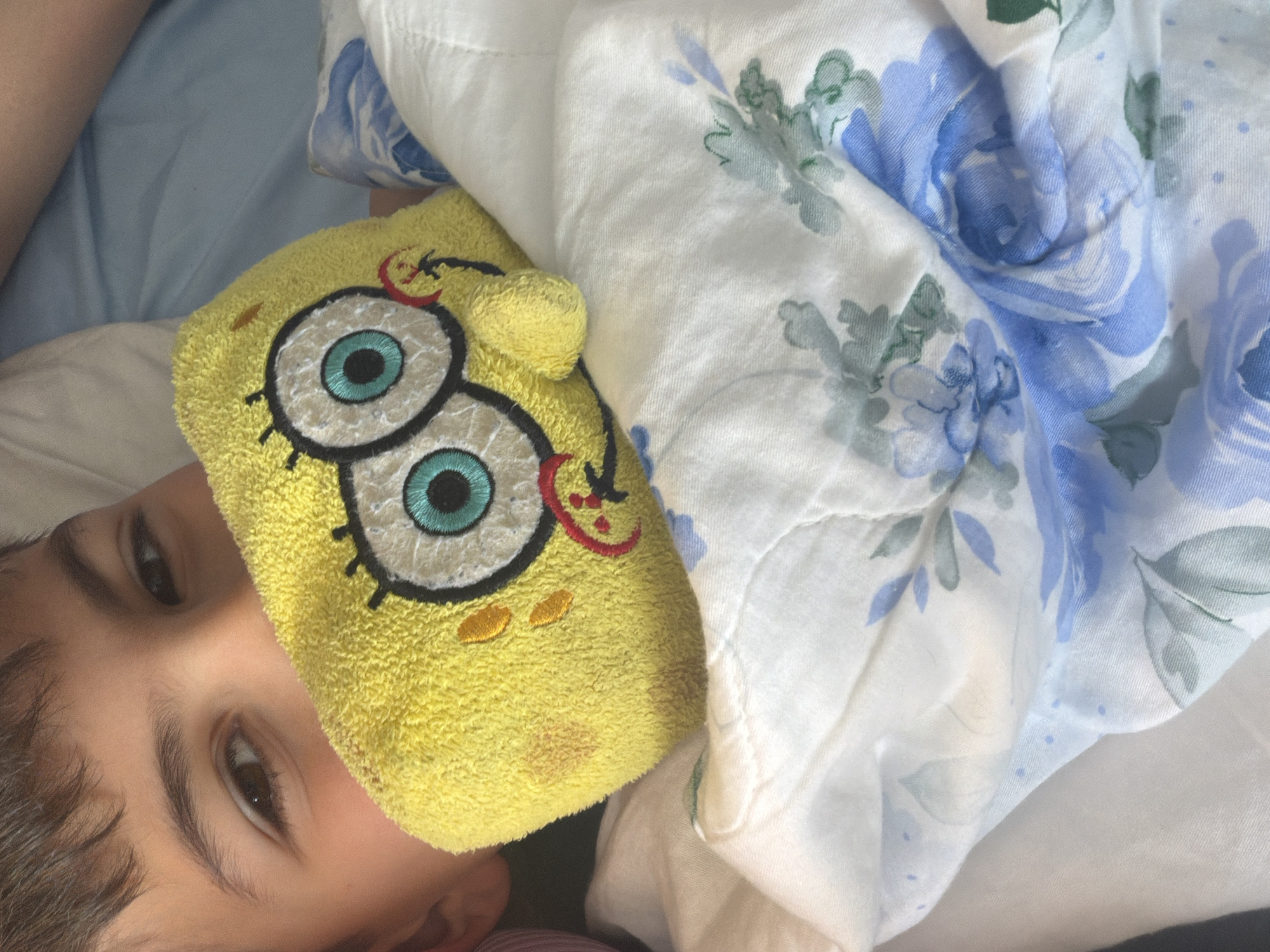 The image shows a close-up, diagonally oriented shot of a young child lying in bed, partially obscured by bedding and a novelty eye mask. The child's face, specifically their right eye, eyebrow, and part of their forehead with dark hair, is visible in the lower left. A vibrant yellow Spongebob Squarepants eye mask covers the central part of the child's face, with one of Spongebob's large, light blue eyes and parts of its embroidered smile and freckles prominently displayed.
The child appears to be resting or waking up, with a serene expression in their visible dark eye. They are partially covered by a white blanket or duvet adorned with a pattern of soft blue roses and green leaves, which extends across the top and right side of the frame. In the upper left background, a plain light blue fabric, likely another sheet or pillowcase, is visible.
The lighting suggests it is daytime, possibly morning, given the context of being in bed with an eye mask. The scene conveys a calm and peaceful atmosphere within an indoor setting, likely a bedroom. While the image itself does not offer specific geographical cues, the provided context places this moment in Budva, Montenegro. There is no visible text in the image.